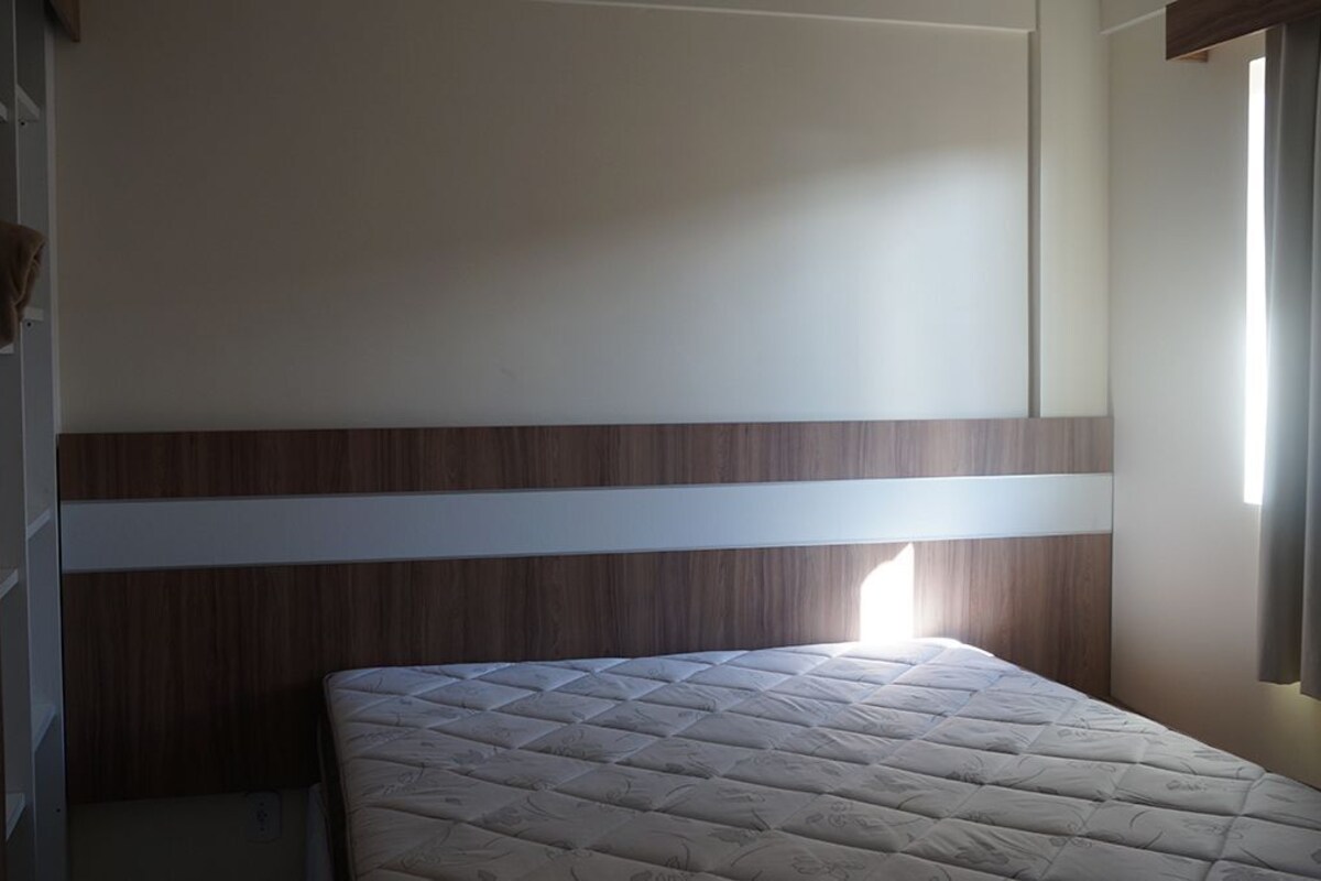 Una habitación con una cama, cabecera de madera, una ventana con cortinas y una puerta.