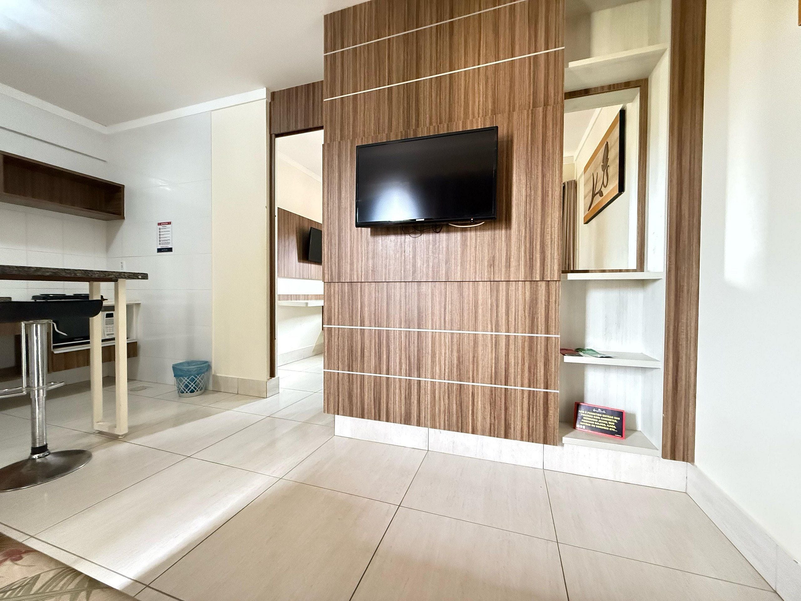 A modern kitchen with a flat-screen TV mounted on a wooden panel wall, a dining area with a table and chairs, and a trash bin.
