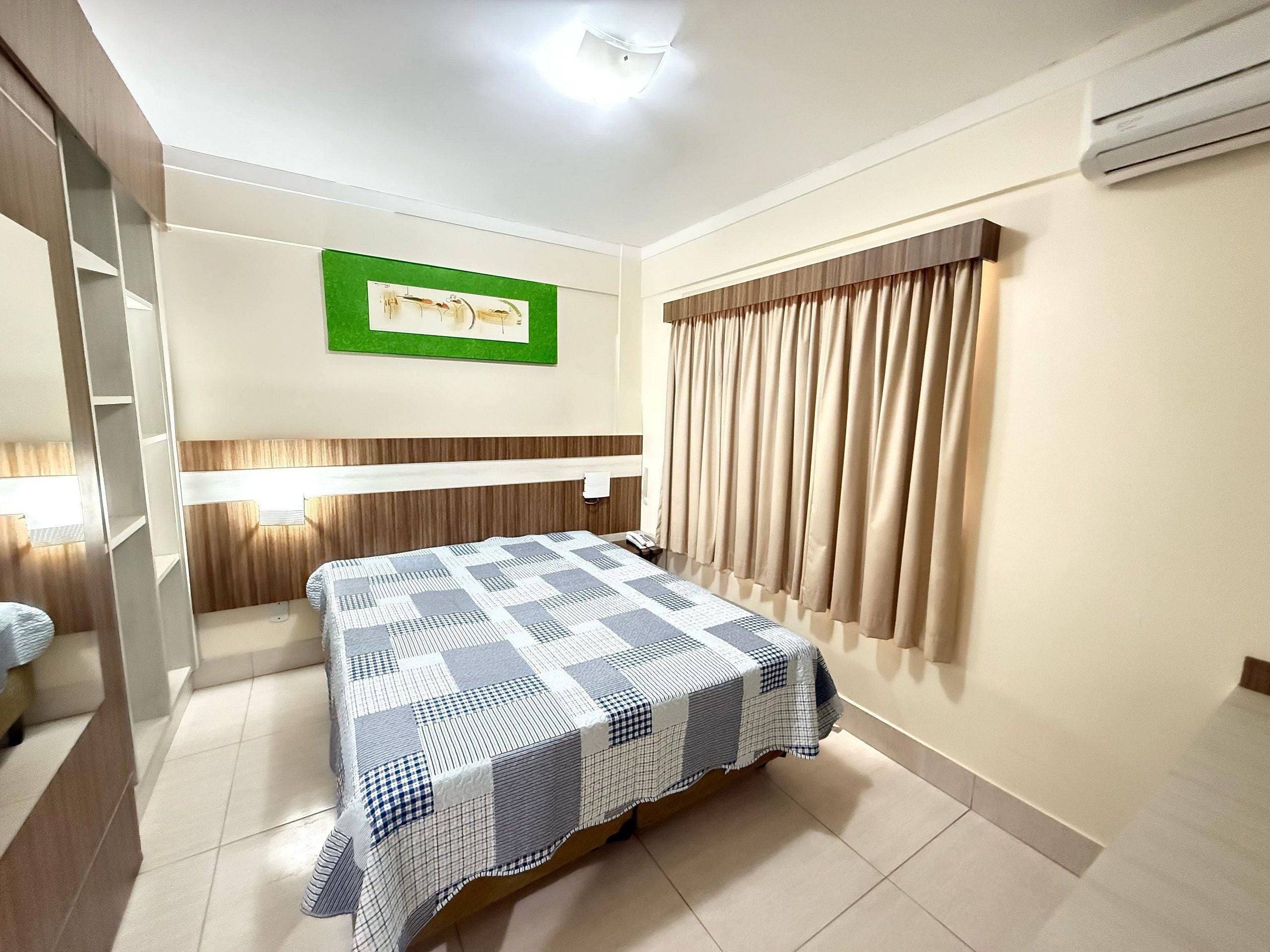 A hotel room with a bed, a nightstand, a mirror, a wall-mounted air conditioner, and a window with curtains.