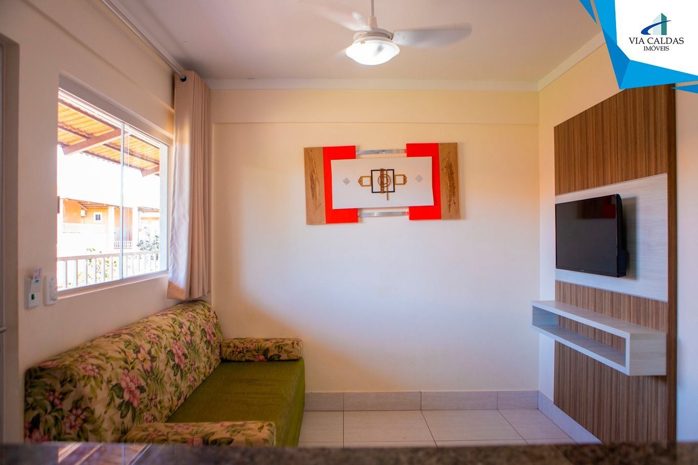 A room with a sofa, a TV mounted on a wooden panel, a ceiling fan, and a window with curtains.