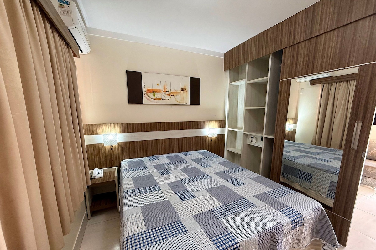 A hotel room with a bed, bedside table, lamp, wall art, wardrobe, and a mirror.