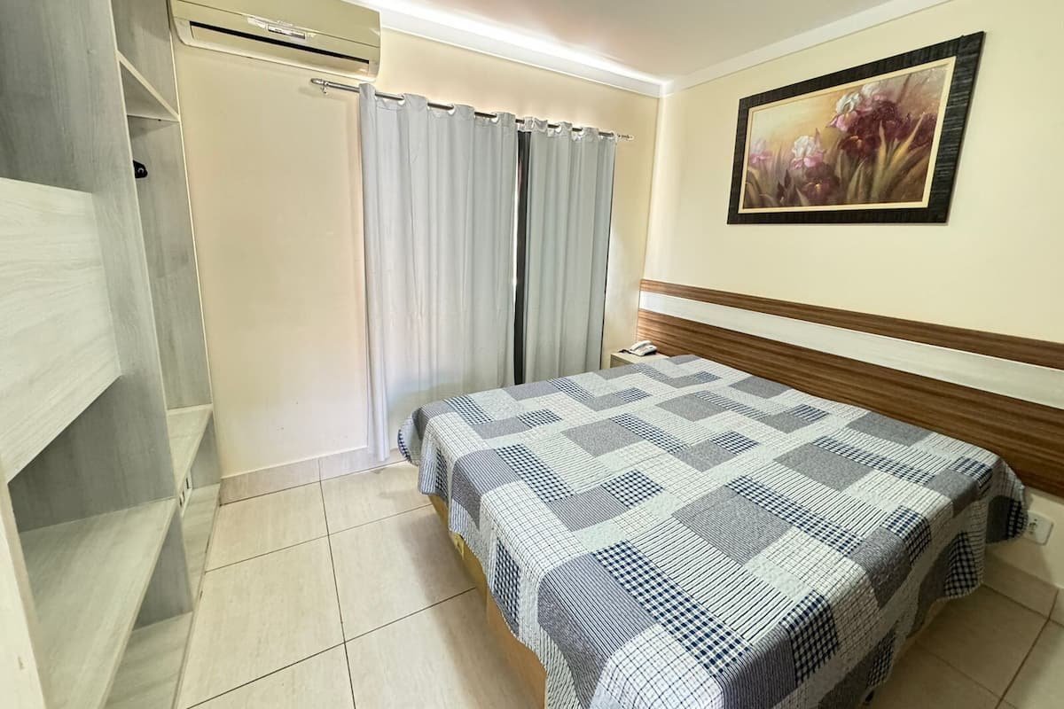 A hotel room with a bed, an air conditioning unit, a curtain, a painting on the wall, and a tiled floor.