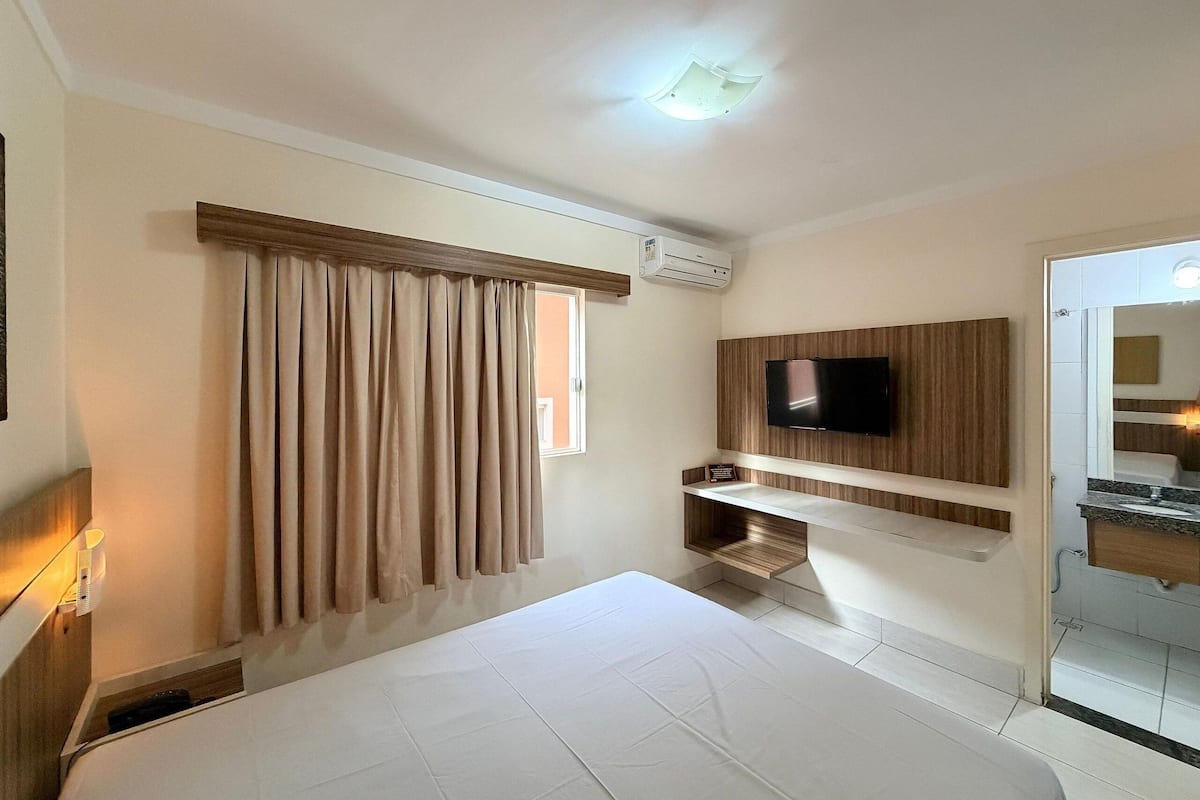 A hotel room with a bed, a television mounted on the wall, a window with curtains, and an air conditioning unit.