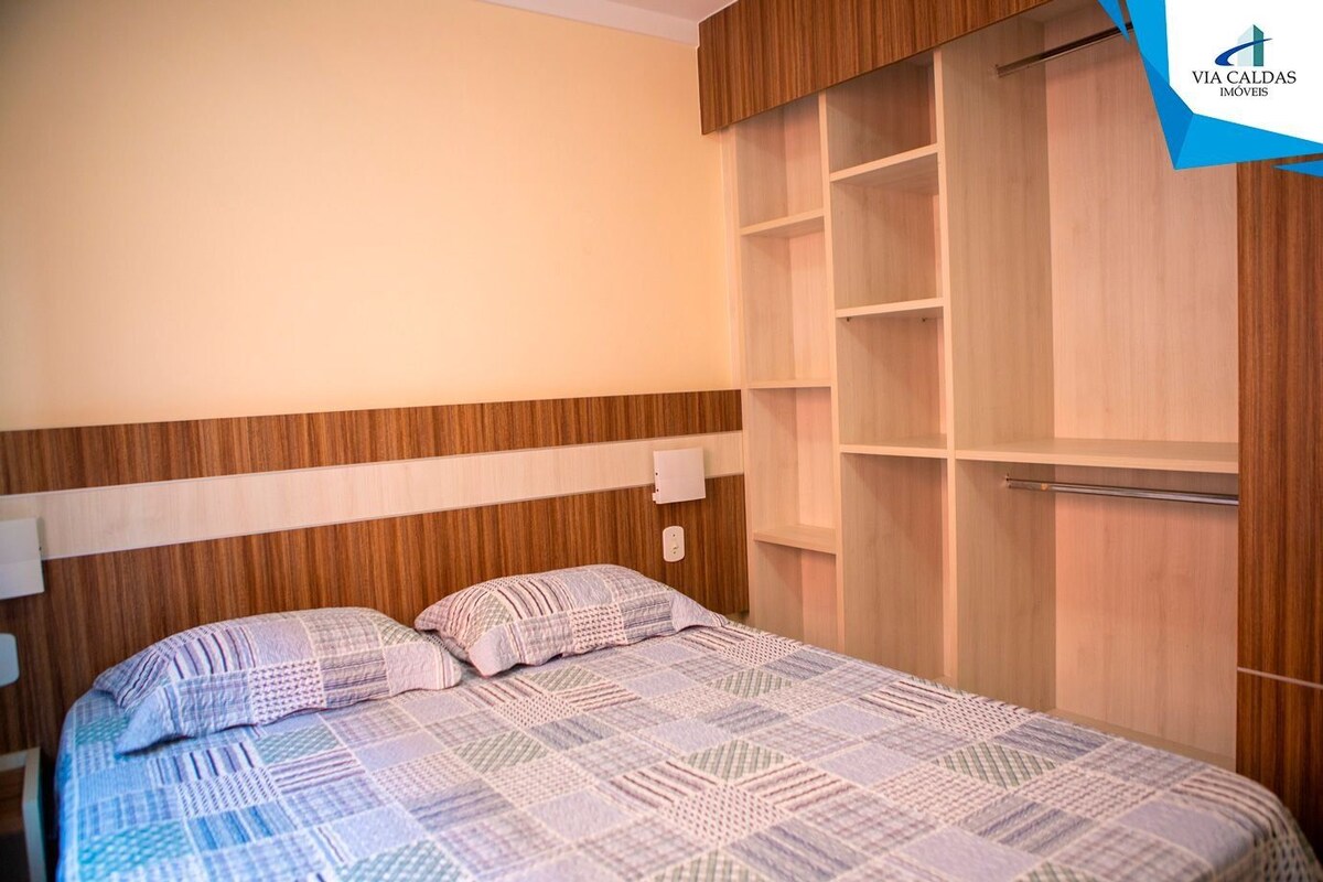A bedroom with a bed, a nightstand, a wooden wardrobe with multiple shelves, and a wall-mounted light.