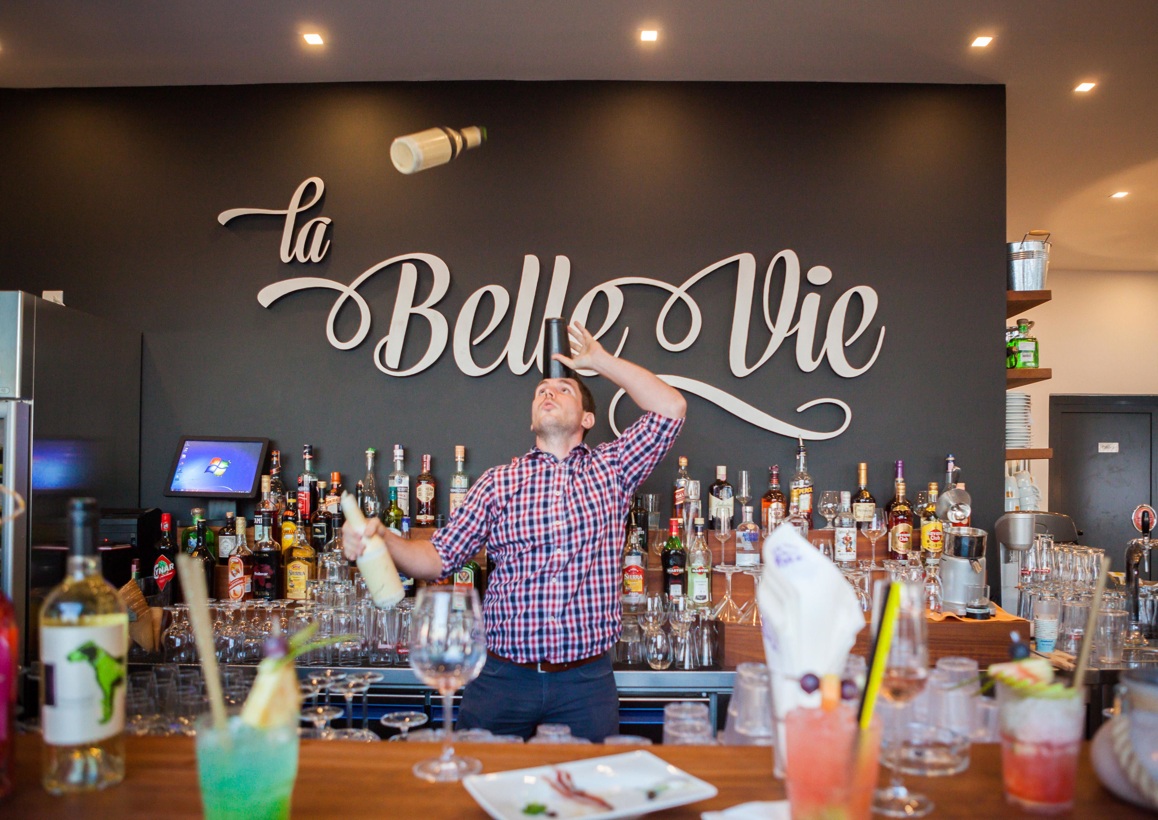 A bartender at a bar with a large sign reading "la Belle Vie".
