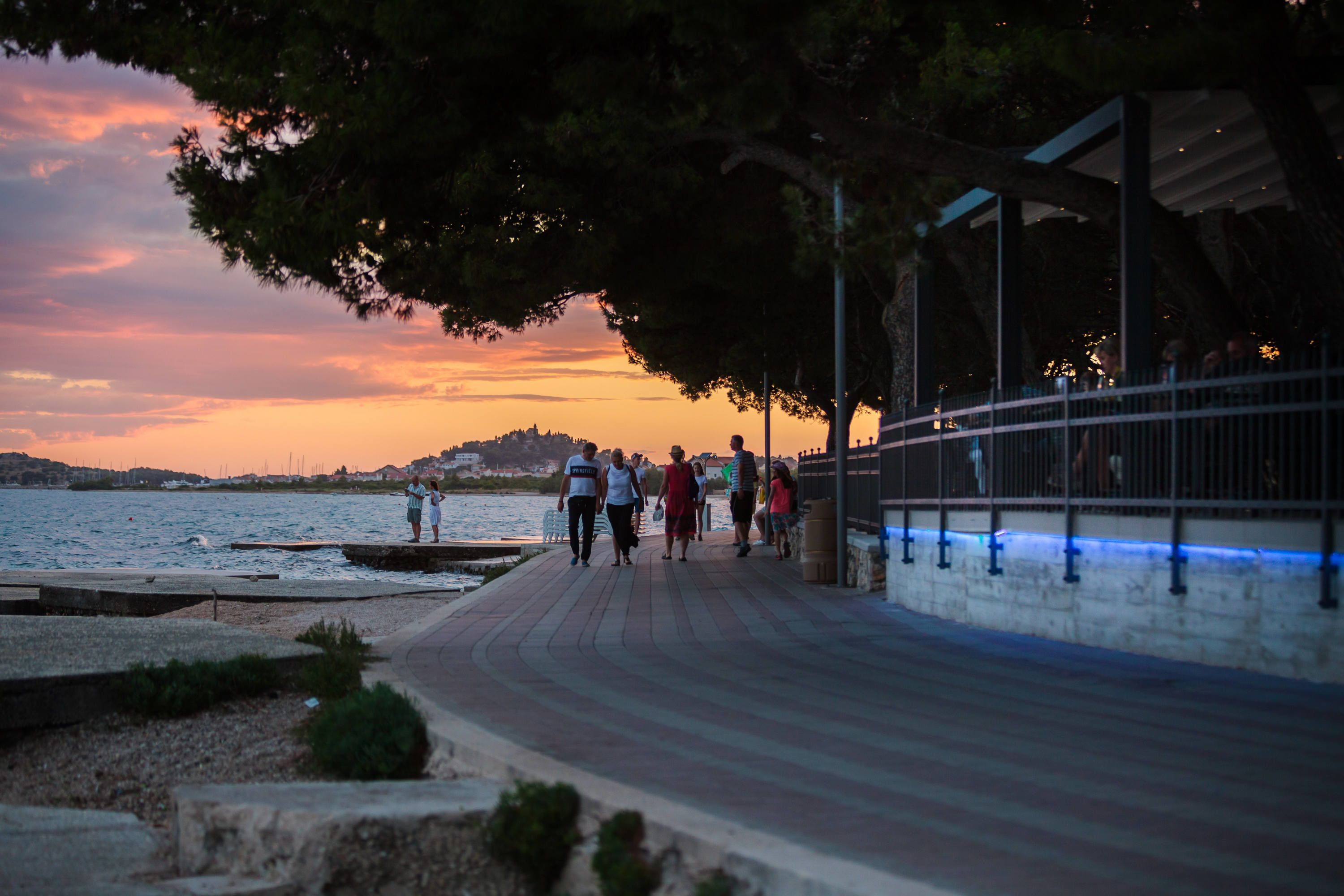 Eine Uferpromenade mit Spaziergängern, einem Steg und Blick auf den Sonnenuntergang.