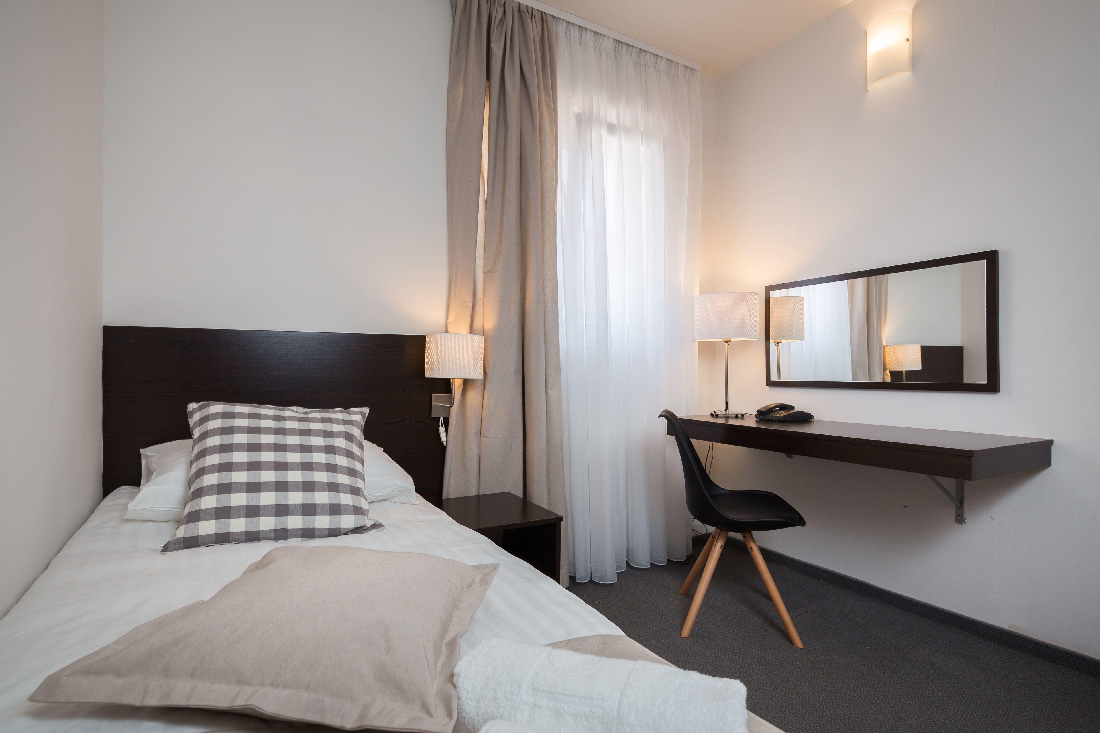 A hotel room with a bed, a desk, a chair, a mirror, and a nightstand with a lamp.