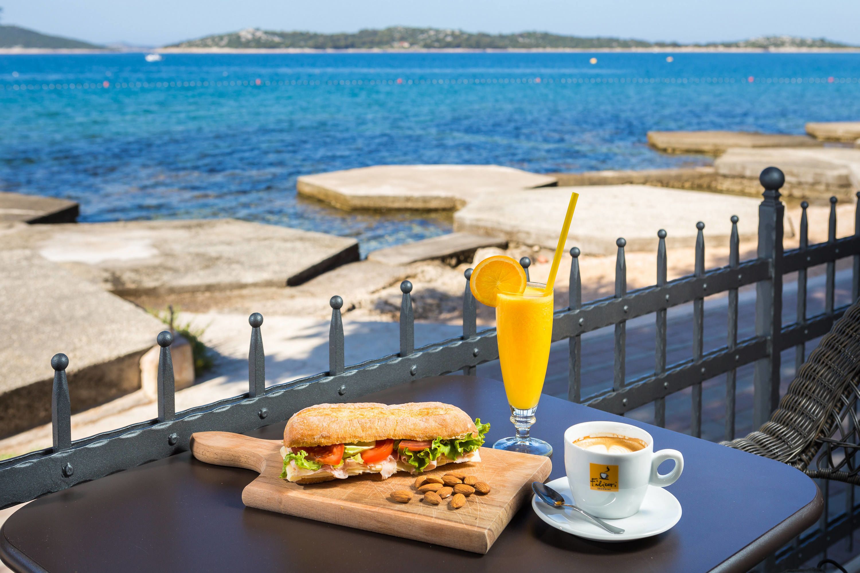 A table with a sandwich, a glass of orange juice, and a cup of coffee, overlooking a body of water.