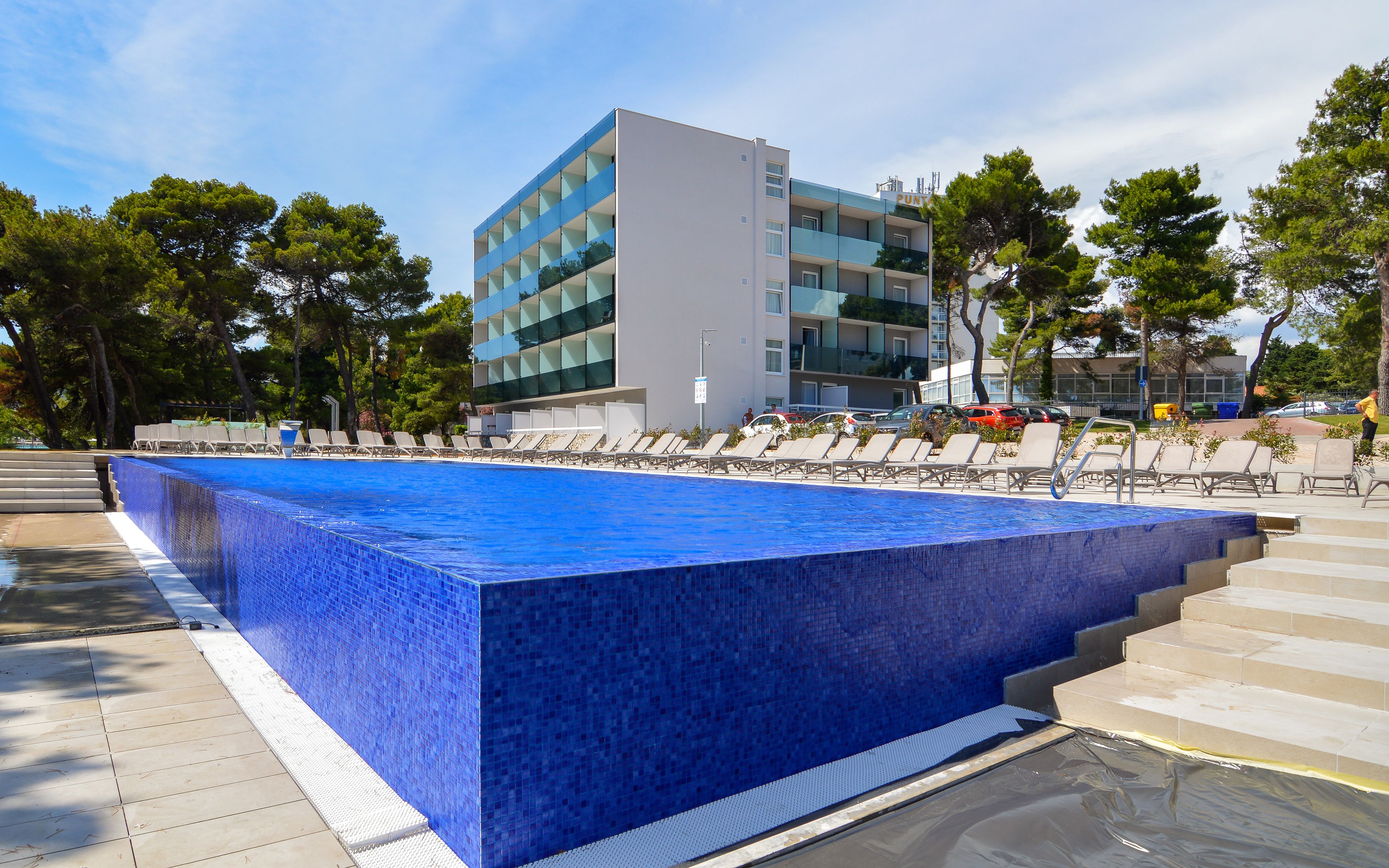 A swimming pool with a blue tiled border, surrounded by lounge chairs and a modern building in the background.