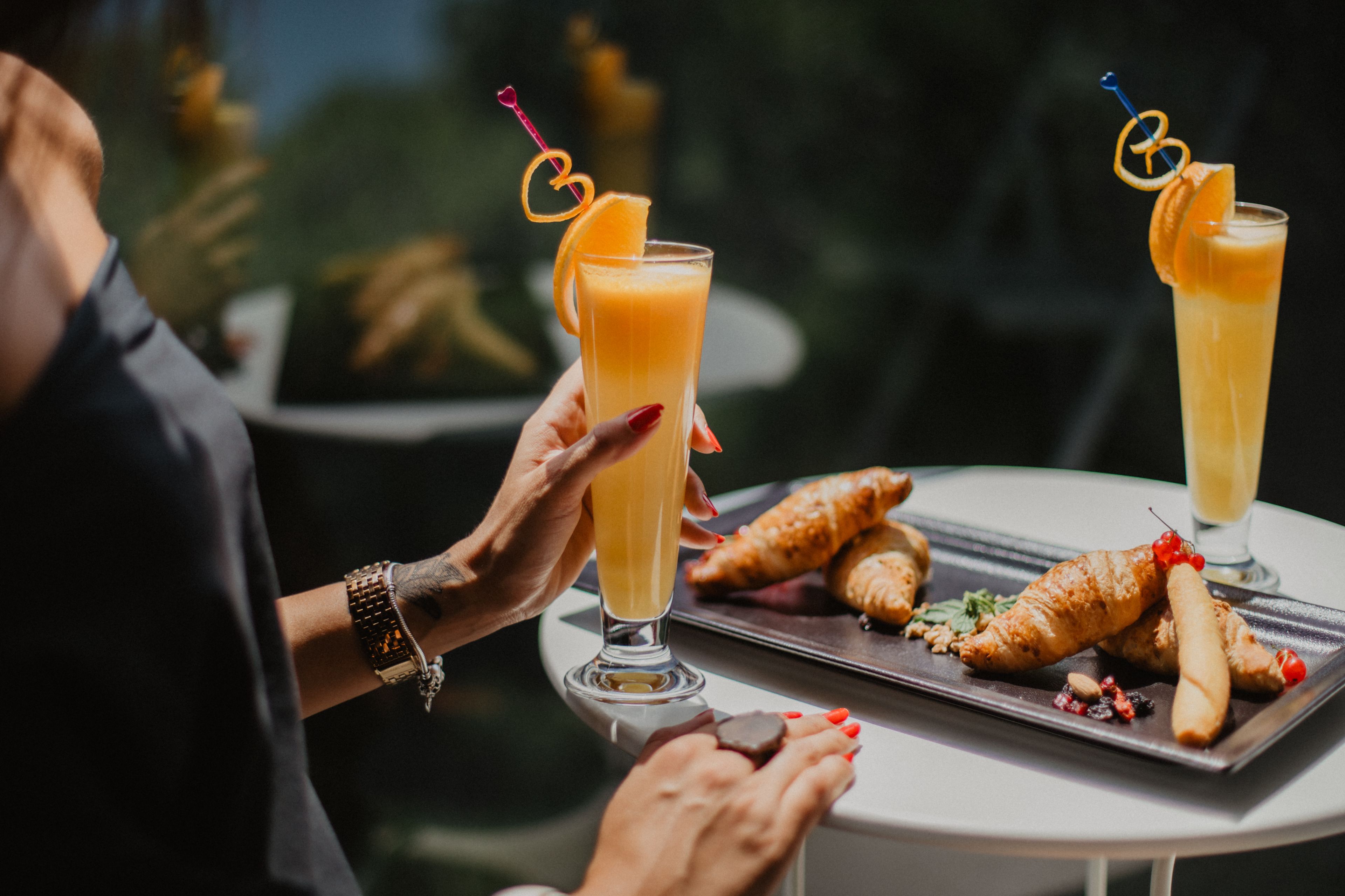 Two people holding tall glasses of orange juice with garnishes, a croissant, and a small pastry on a tray.