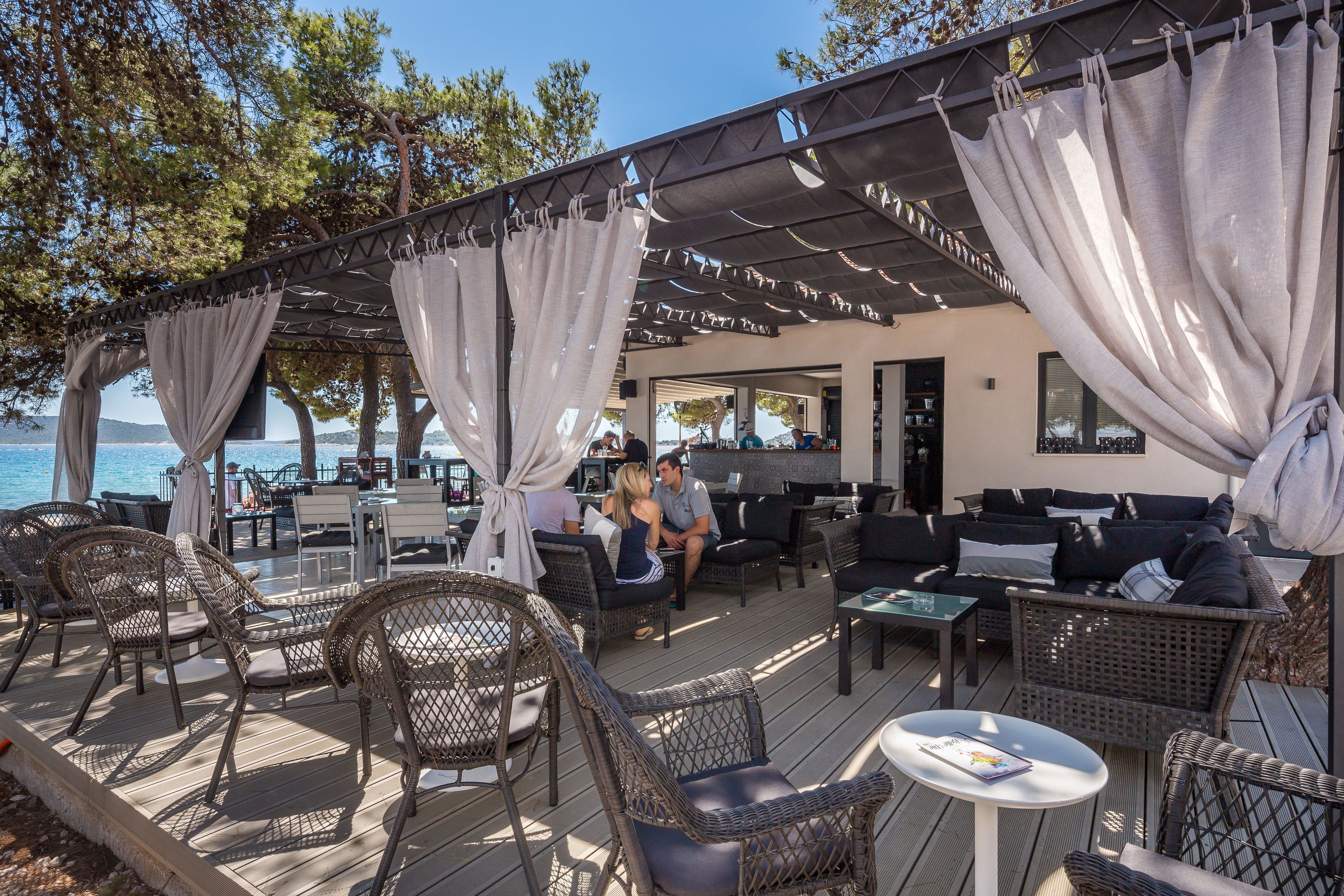 An outdoor seating area with wicker furniture and a covered section with curtains, overlooking a body of water.