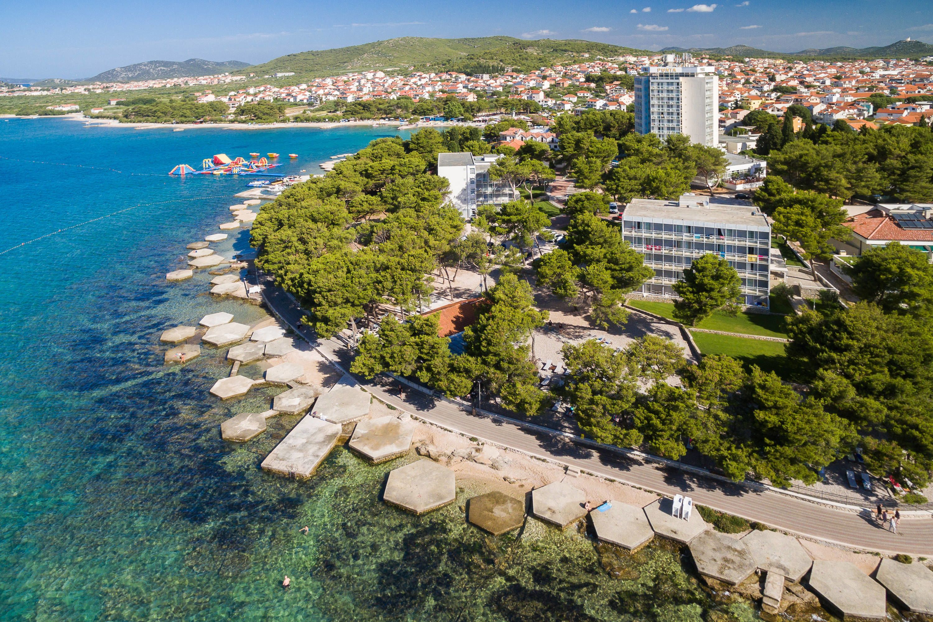 An aerial view of a coastal town with clear blue waters, a promenade lined with hexagonal stone slabs, and a mix of residential and commercial buildings.