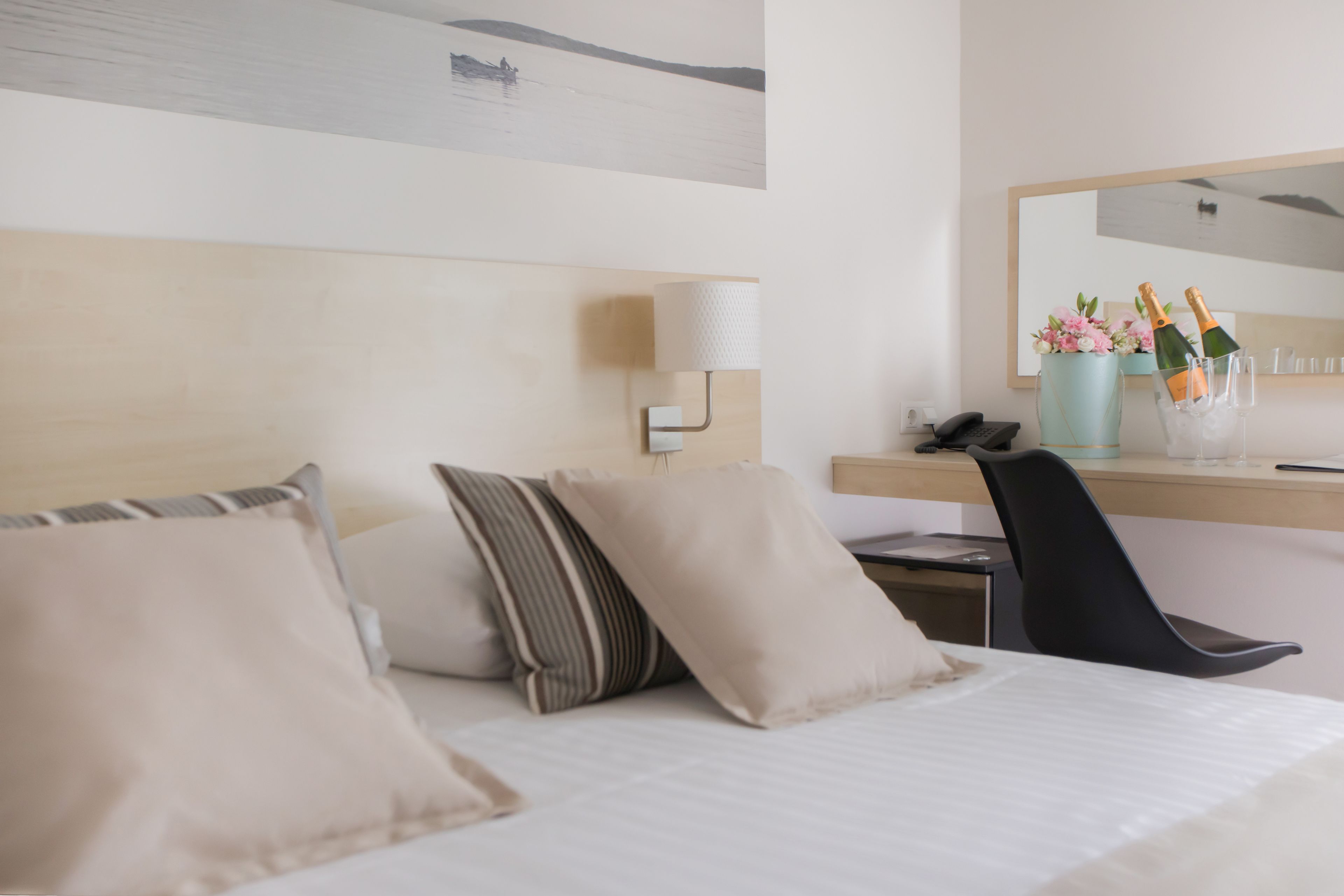 A hotel room with a bed, pillows, a desk with a lamp, a chair, and a mirror. There are two bottles of champagne in a glass container on the desk.
