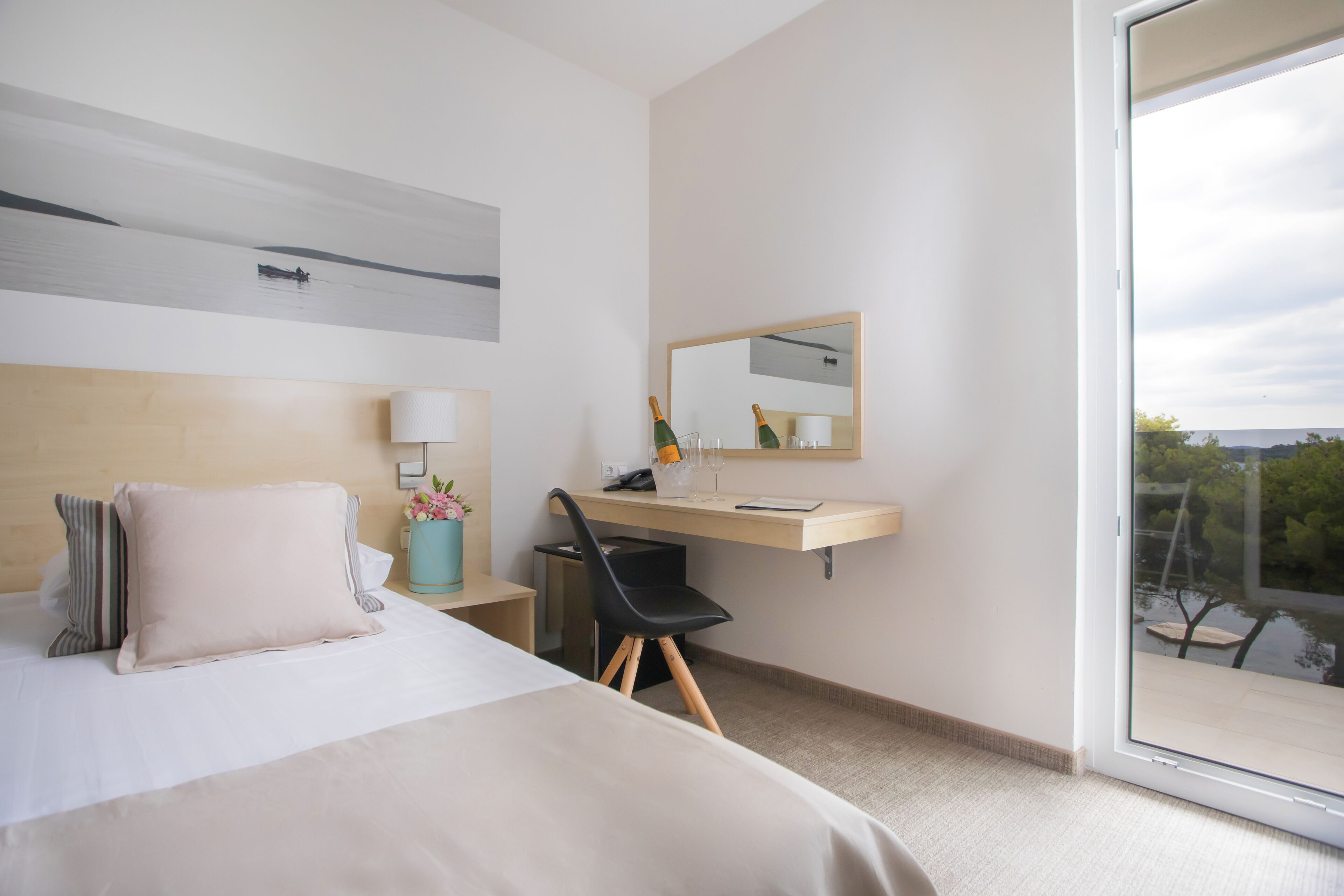A modern hotel room with a bed, a desk, a chair, a mirror, and a view of the outdoors.
