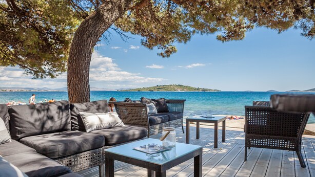 Terraza exterior con sofá, sillas y una mesa, con vista a una playa y al agua.