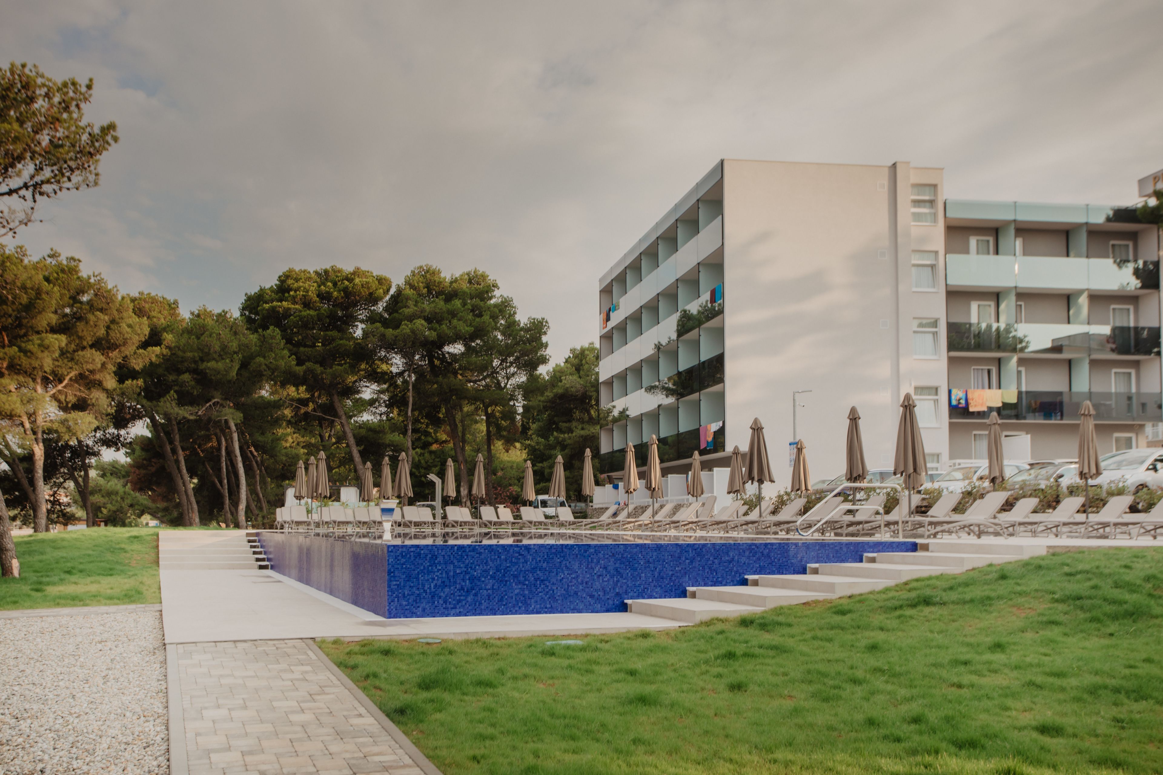 A modern building with a pool area, including lounge chairs and umbrellas, surrounded by trees.