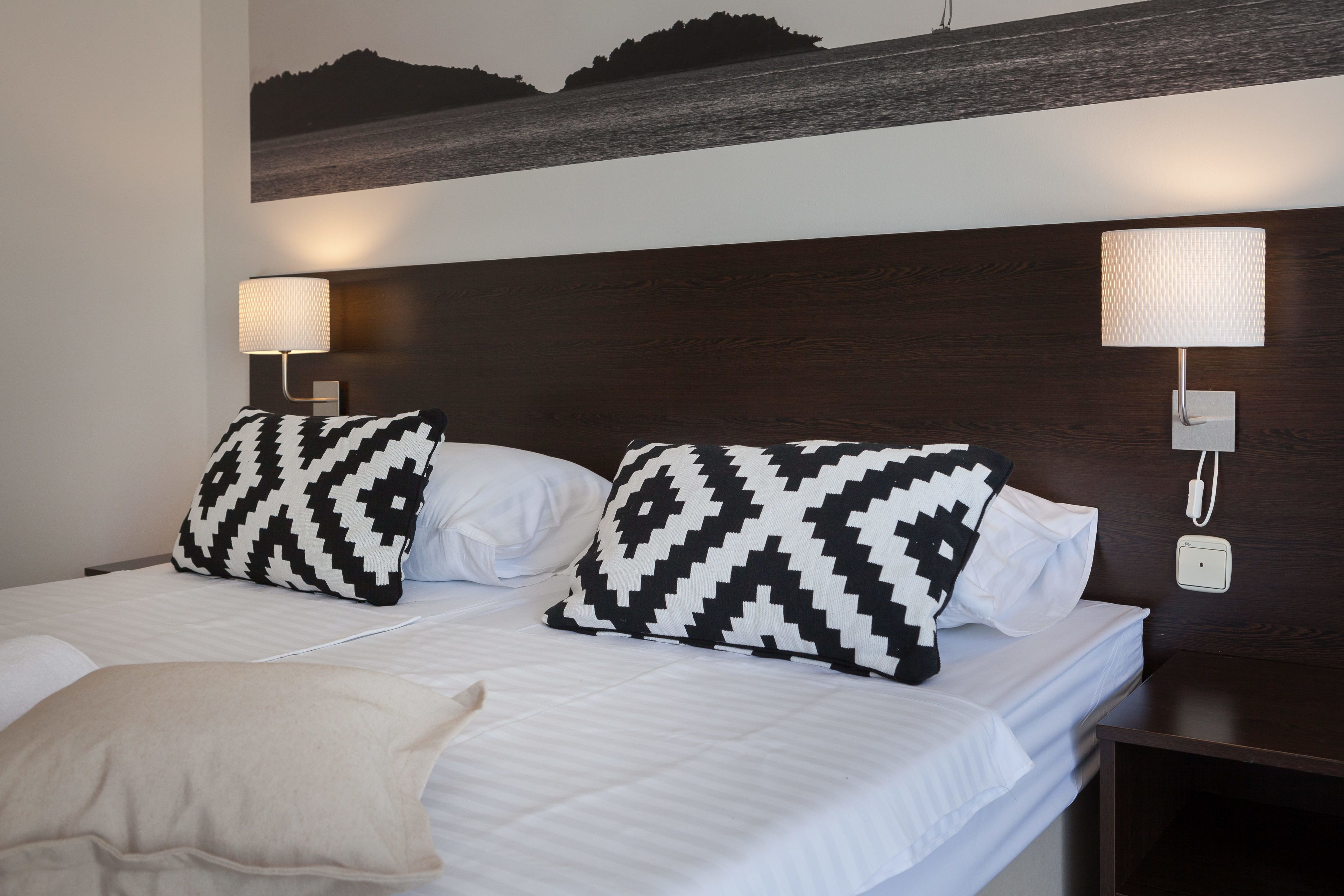 A hotel room with a bed, two patterned pillows, a bedside lamp, and a wall-mounted phone.