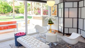 A modern living room with a white sofa, two white chairs, a round ottoman, a rocking chair, a small table with a plant, and a large window with a view of a garden. - Nestled in the Cove (Rancho Mirage)