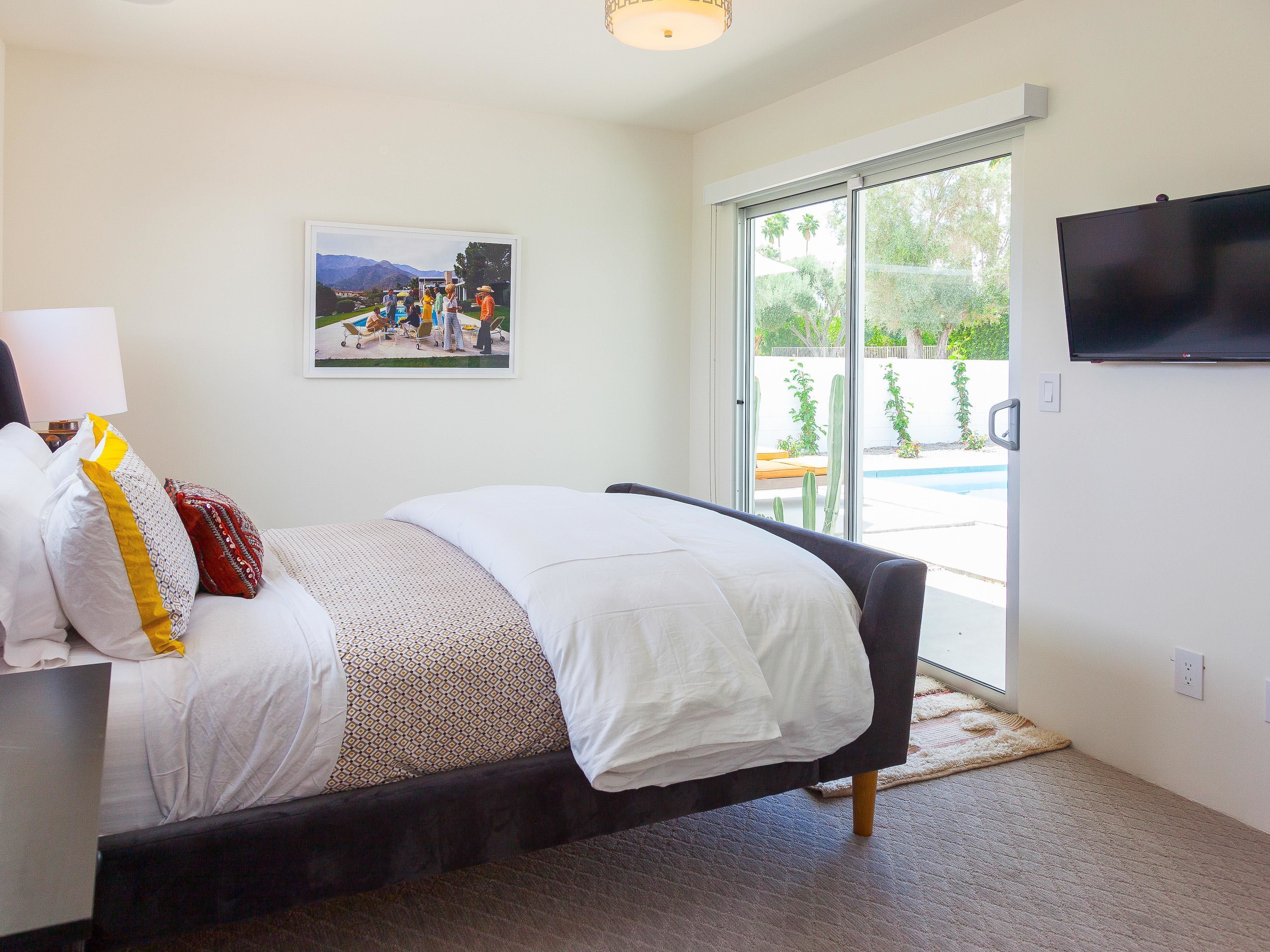 A bedroom with a bed, a TV, a painting, and a view of a pool.