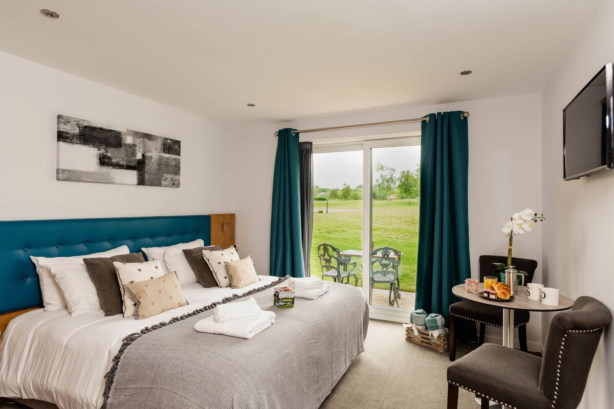 A bedroom with a bed, a bedside table with a flower vase, and a view of the outdoors.