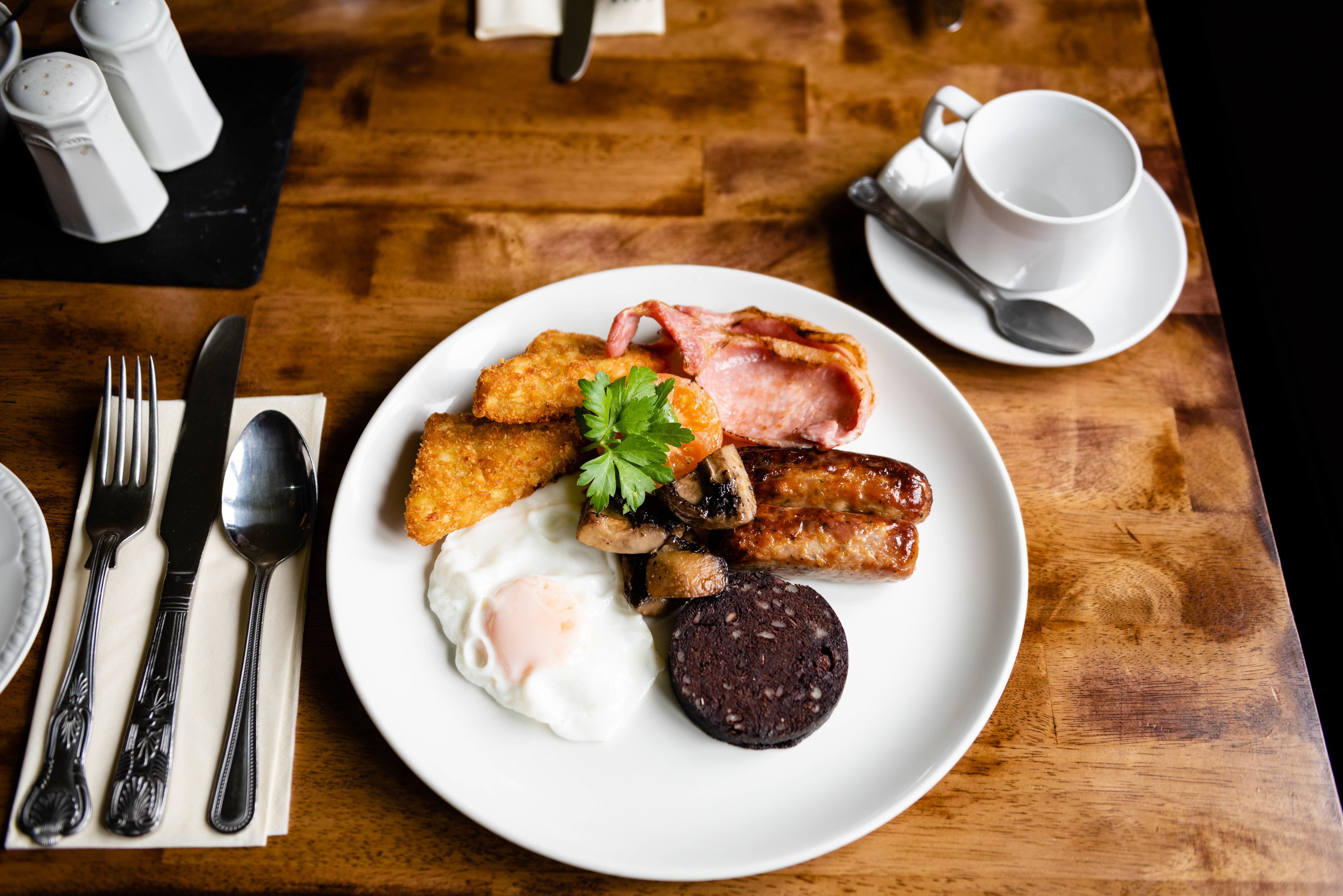 A breakfast plate with eggs, bacon, sausages, and a black pudding, served on a wooden table with a white cup and saucer, and cutlery.