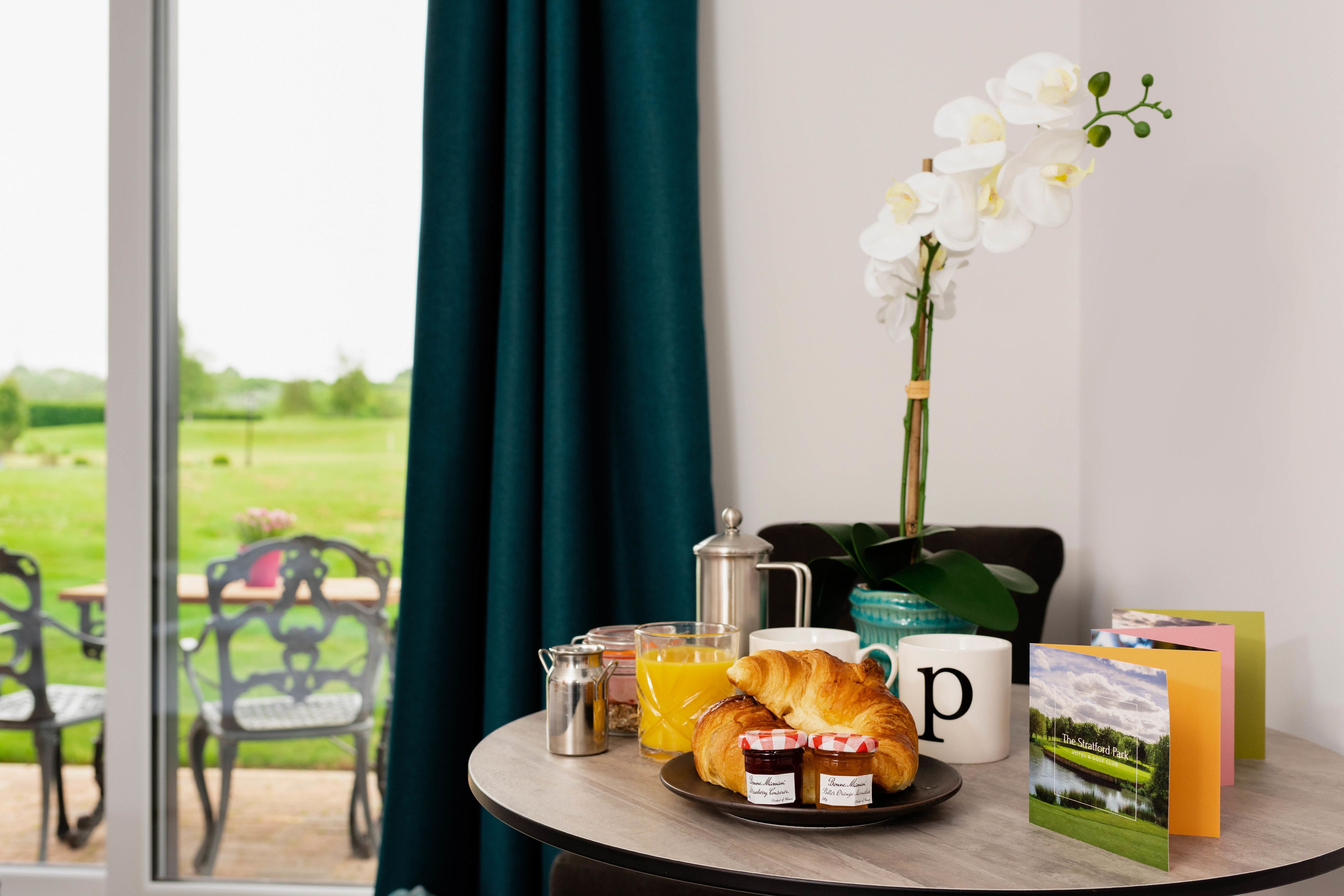 Un desayuno con croissants, mermelada, jugo de naranja y una cafetera sobre una mesa junto a una ventana con vistas a un paisaje verde.