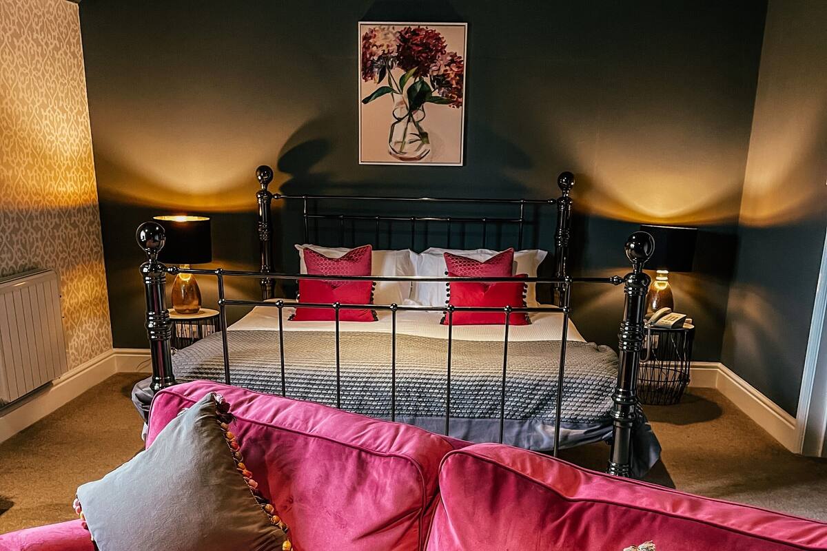 A bedroom with a bed, a red and pink sofa, a painting on the wall, and a lamp.
