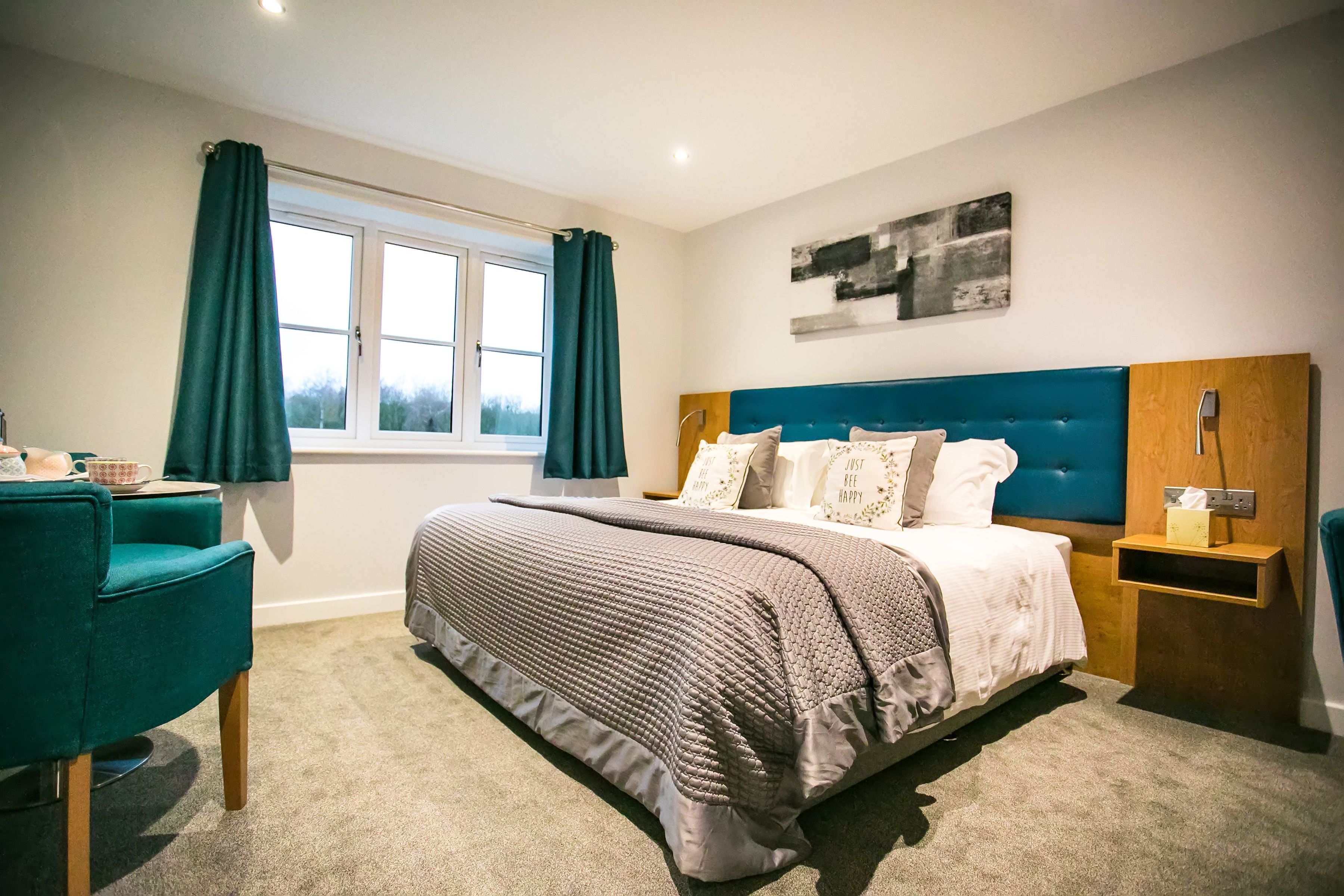 A bedroom with a large bed, a bedside table, a chair, a window with curtains, and a wall-mounted artwork.