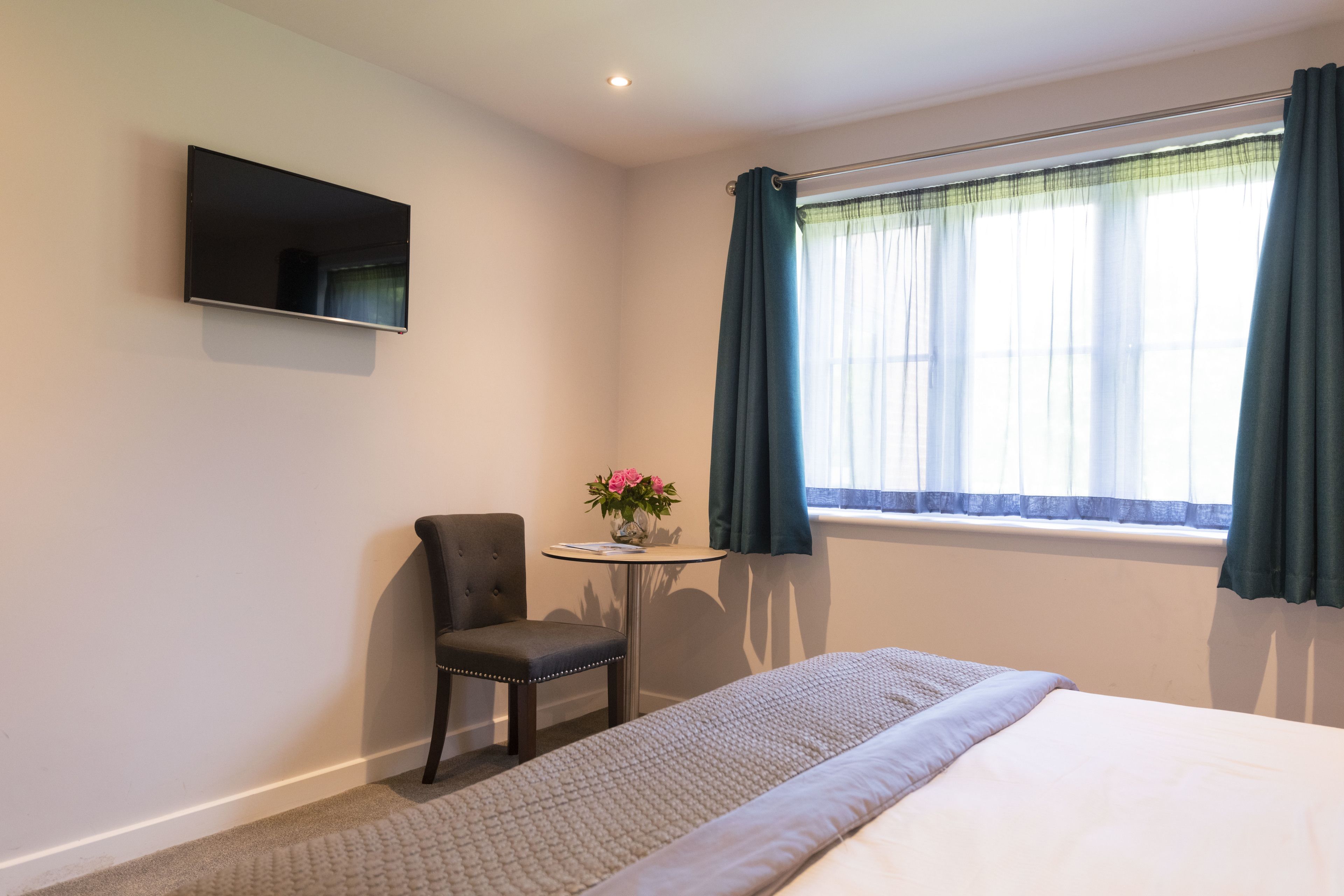 Een hotelkamer met een bed, een stoel, een klein tafeltje met een bloevas en een aan de muur bevestigde televisie.