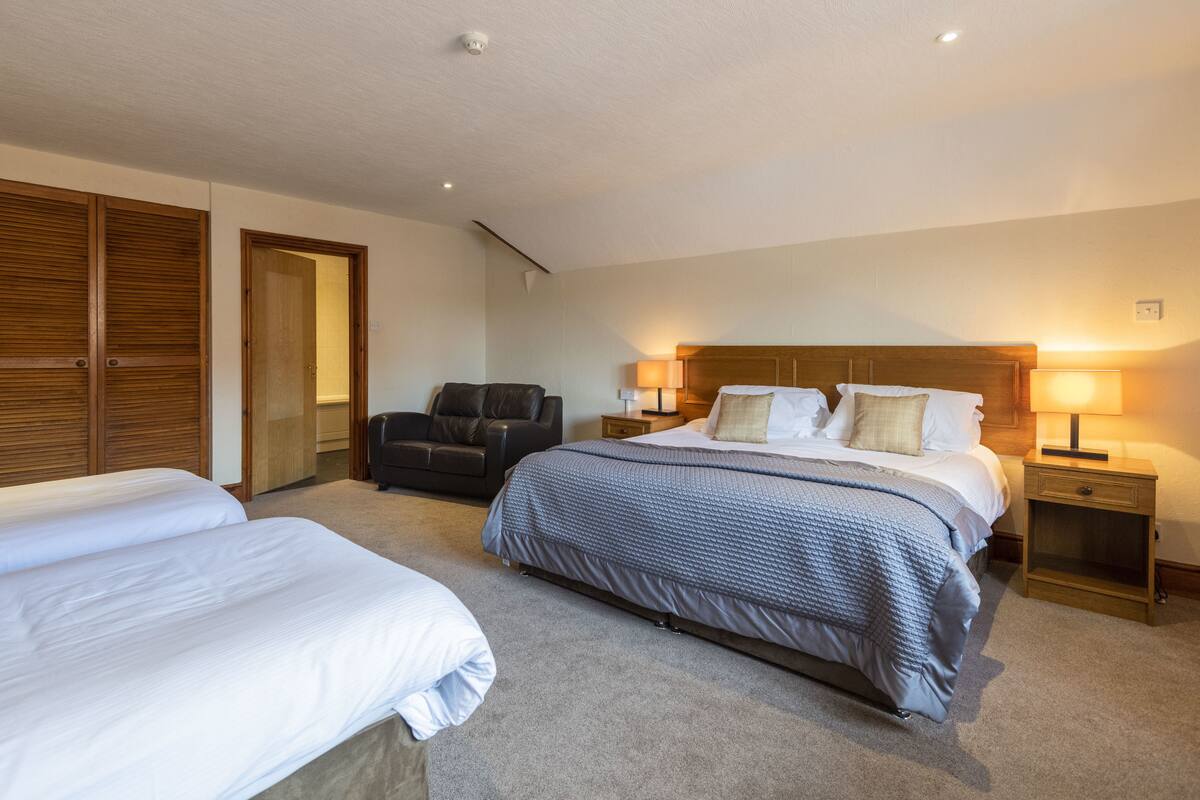 A hotel room with two beds, a sofa, a wooden wardrobe, and a nightstand with a lamp.