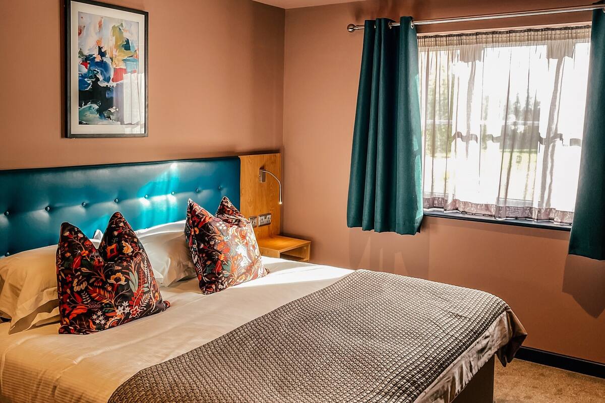 A hotel room with a large bed, a headboard, a bedside table, a nightstand, a window with curtains, and a ceiling light.