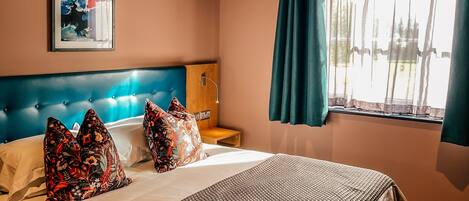 Una habitación de hotel con una cama grande, un cabecero, una mesita de noche y cortinas en la ventana, además de una lámpara en el techo.