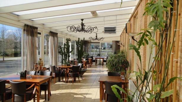 A restaurant with wooden tables and chairs, large windows, and potted plants. - De Wapser Herberg (Wapse)