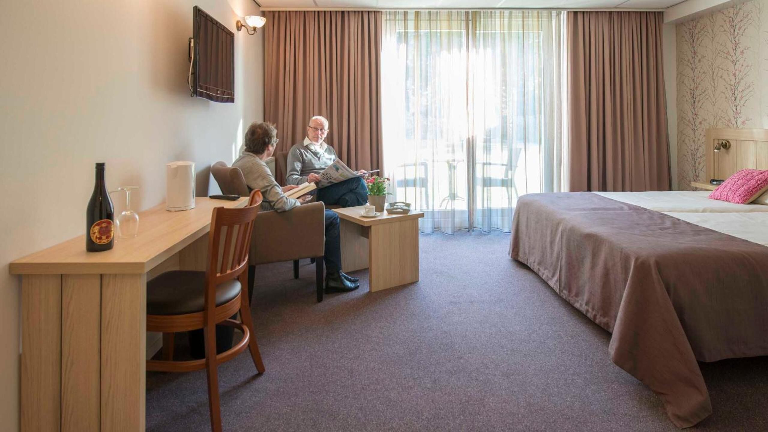 A hotel room with a bed, a desk, and two chairs. There is a bottle and a glass on the desk.