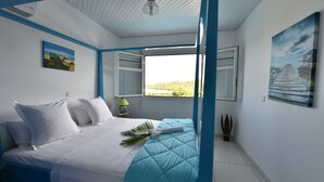 A bedroom with a bed, white bedding, a window with an open shutter, a painting on the wall, and a ceiling fan. - Makare Lagoon Club (Le Vauclin)