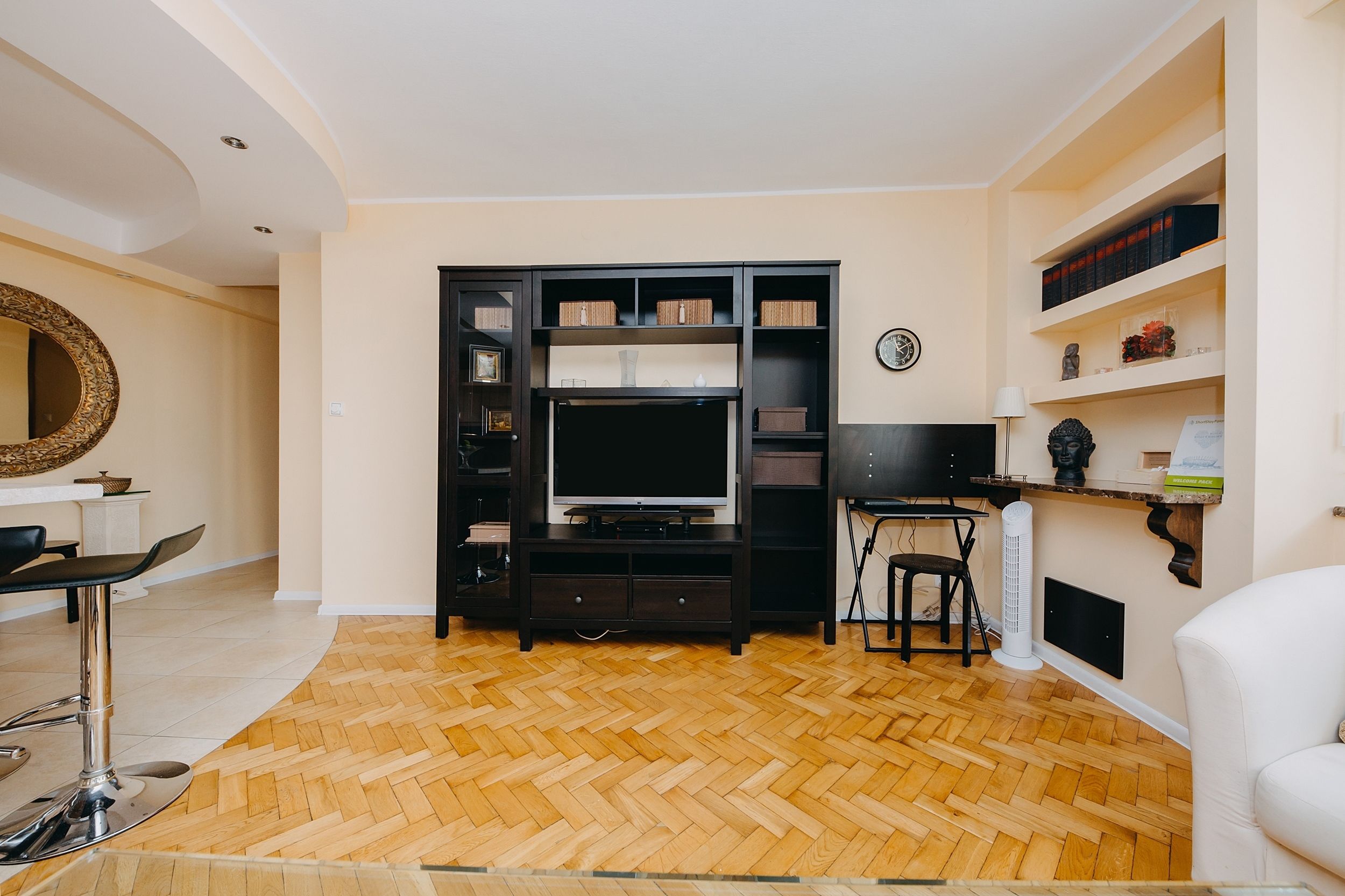 A living room with a herringbone wood floor, a black entertainment unit with a TV, a white sofa, and a small table with a chair.