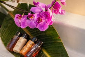 Une branche d’orchidée violette surplombe un évier blanc sur lequel sont posées quatre bouteilles de liquide.