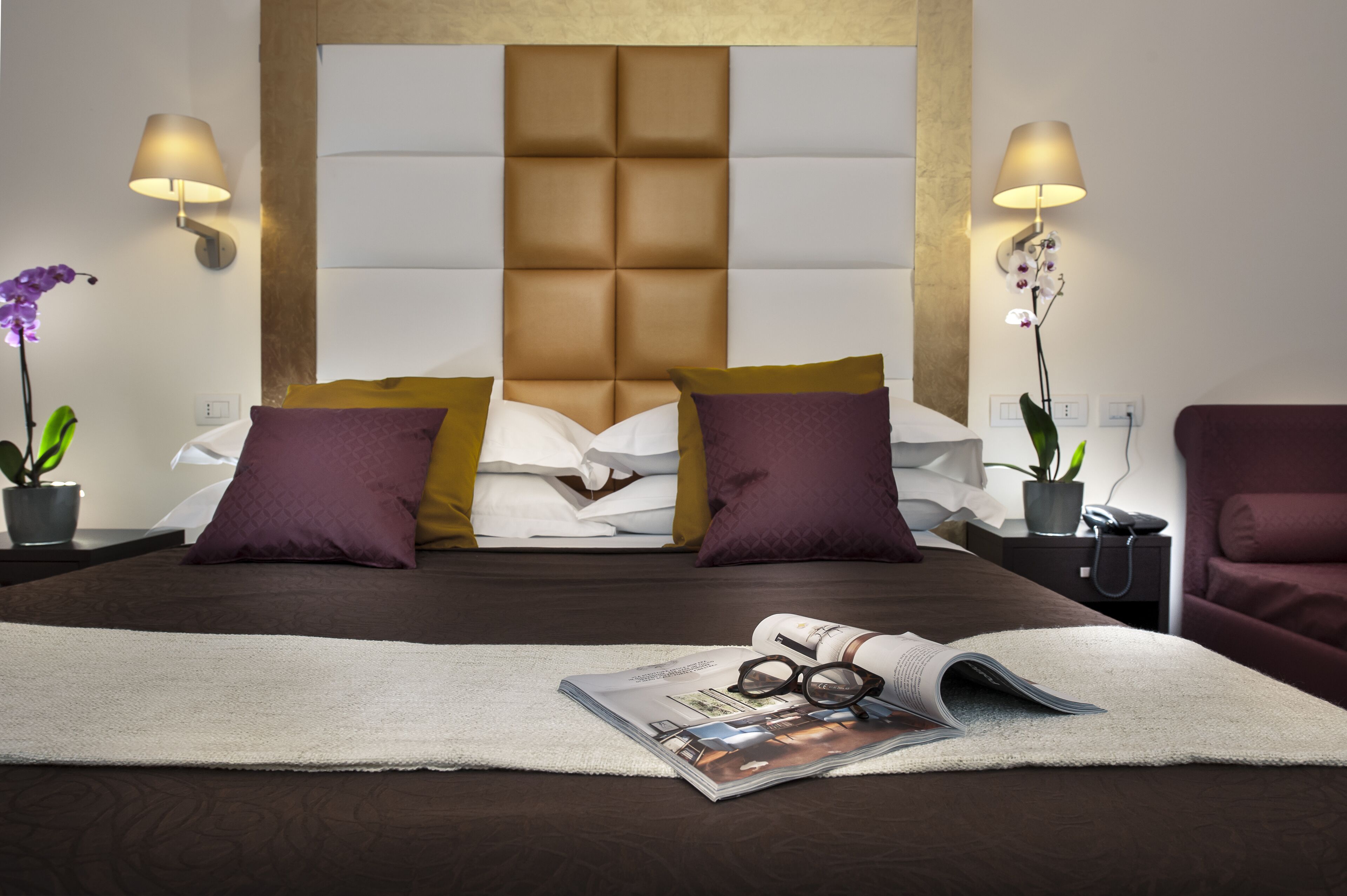 A neatly made bed with a headboard, two bedside lamps, and a magazine with glasses on it.