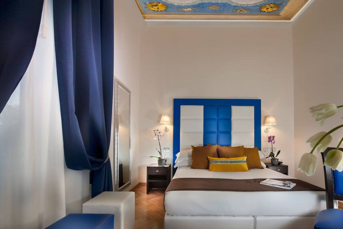 A hotel room with a large bed, a blue headboard, two bedside tables with lamps, and a ceiling with a decorative design.