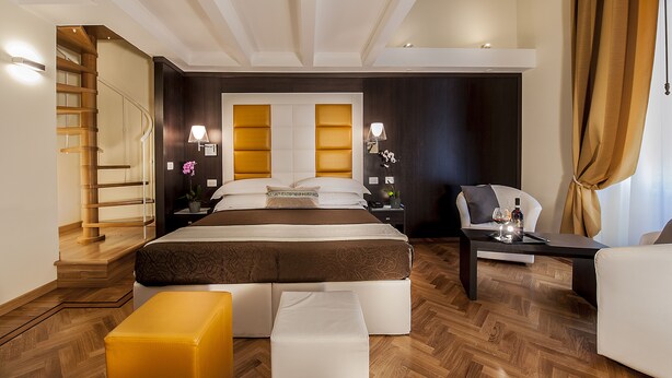 A modern hotel room with a large bed, a sofa, a small table, and a chair. There is a staircase leading to another level. - DVE Suite Rome (Rome)