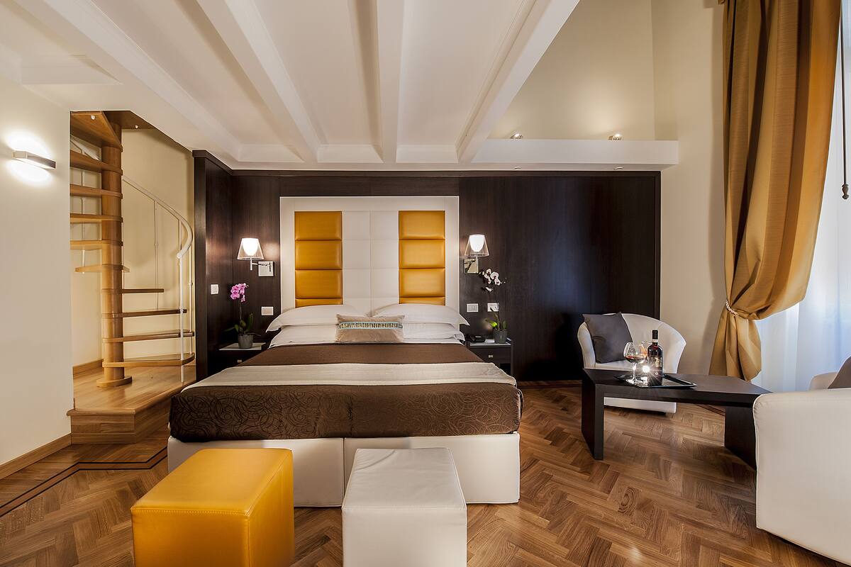 A modern hotel room with a large bed, a sofa, a small table, and a chair. There is a staircase leading to another level.