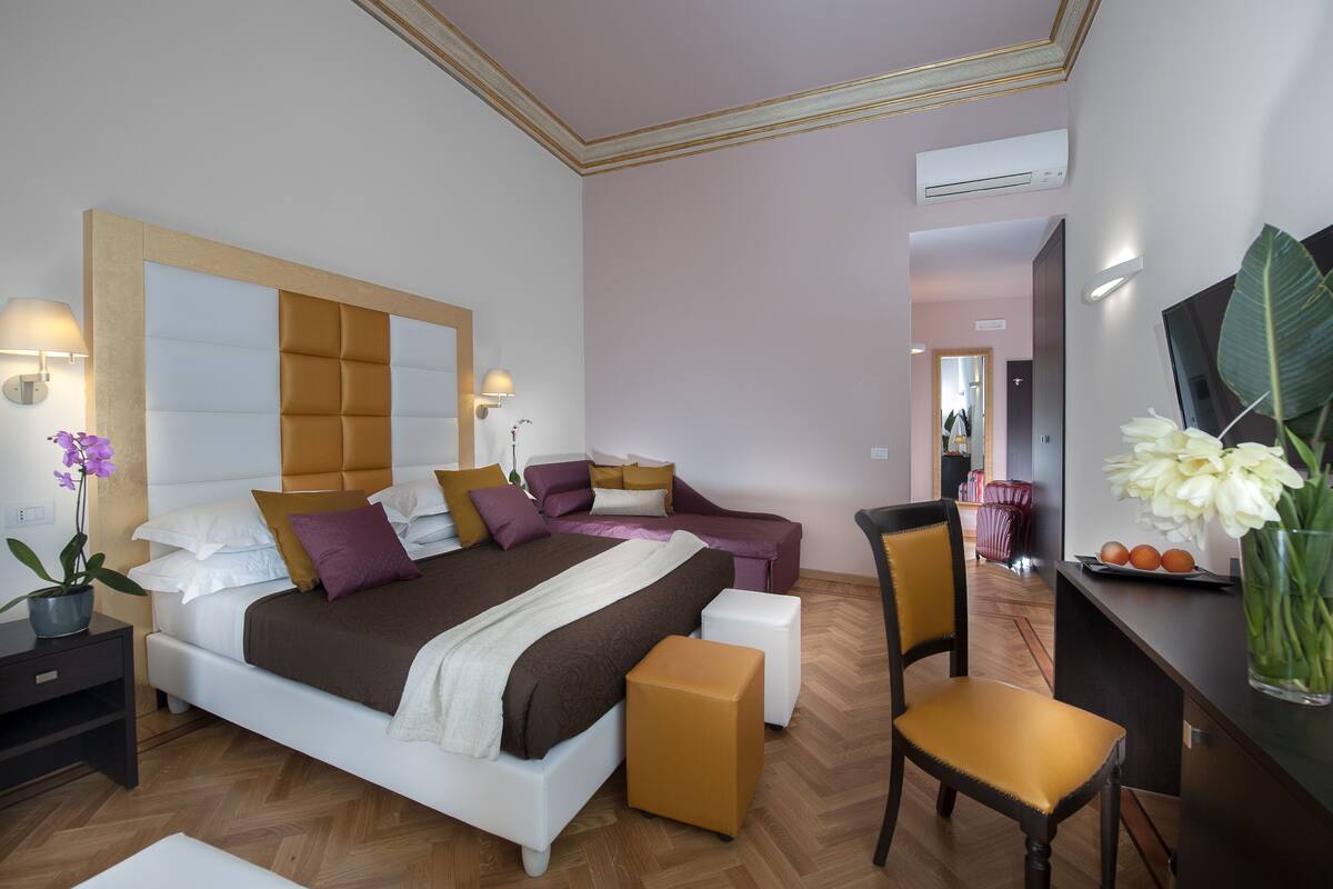 A modern hotel room with a large bed, a sofa, a desk, and a chair. There is a potted plant and a vase with flowers on the desk.