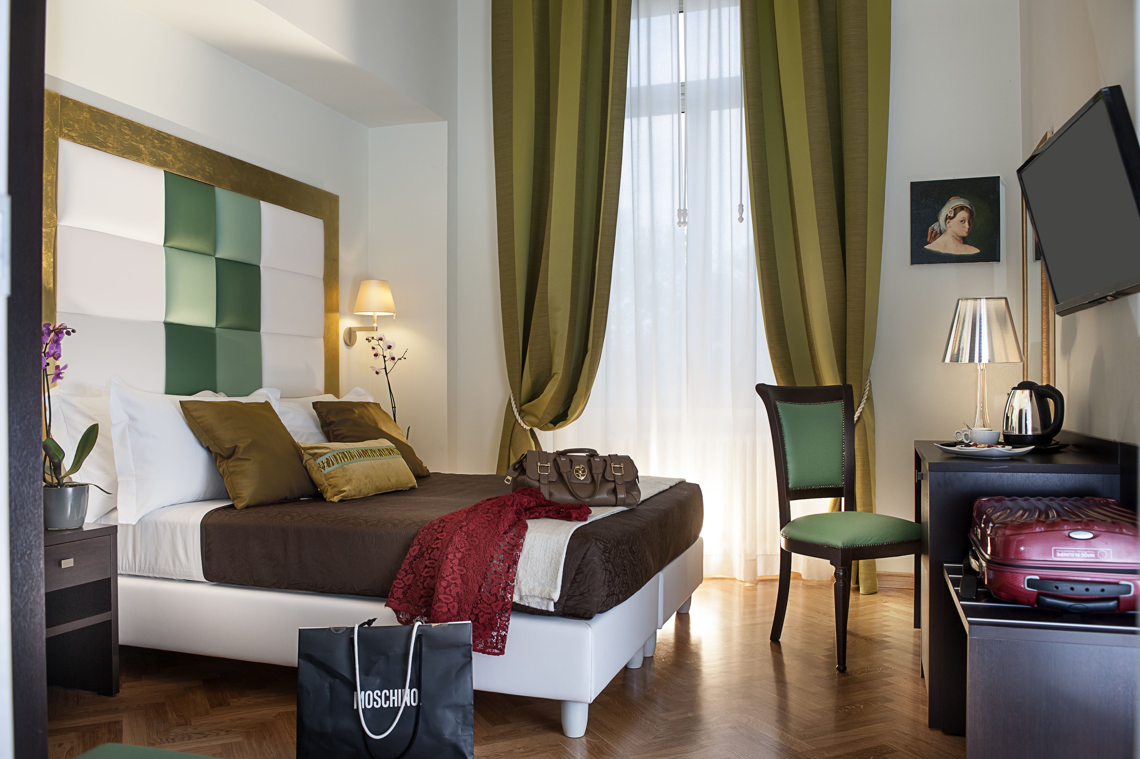 A hotel room with a bed, a chair, a desk with a laptop, a suitcase, and a television.