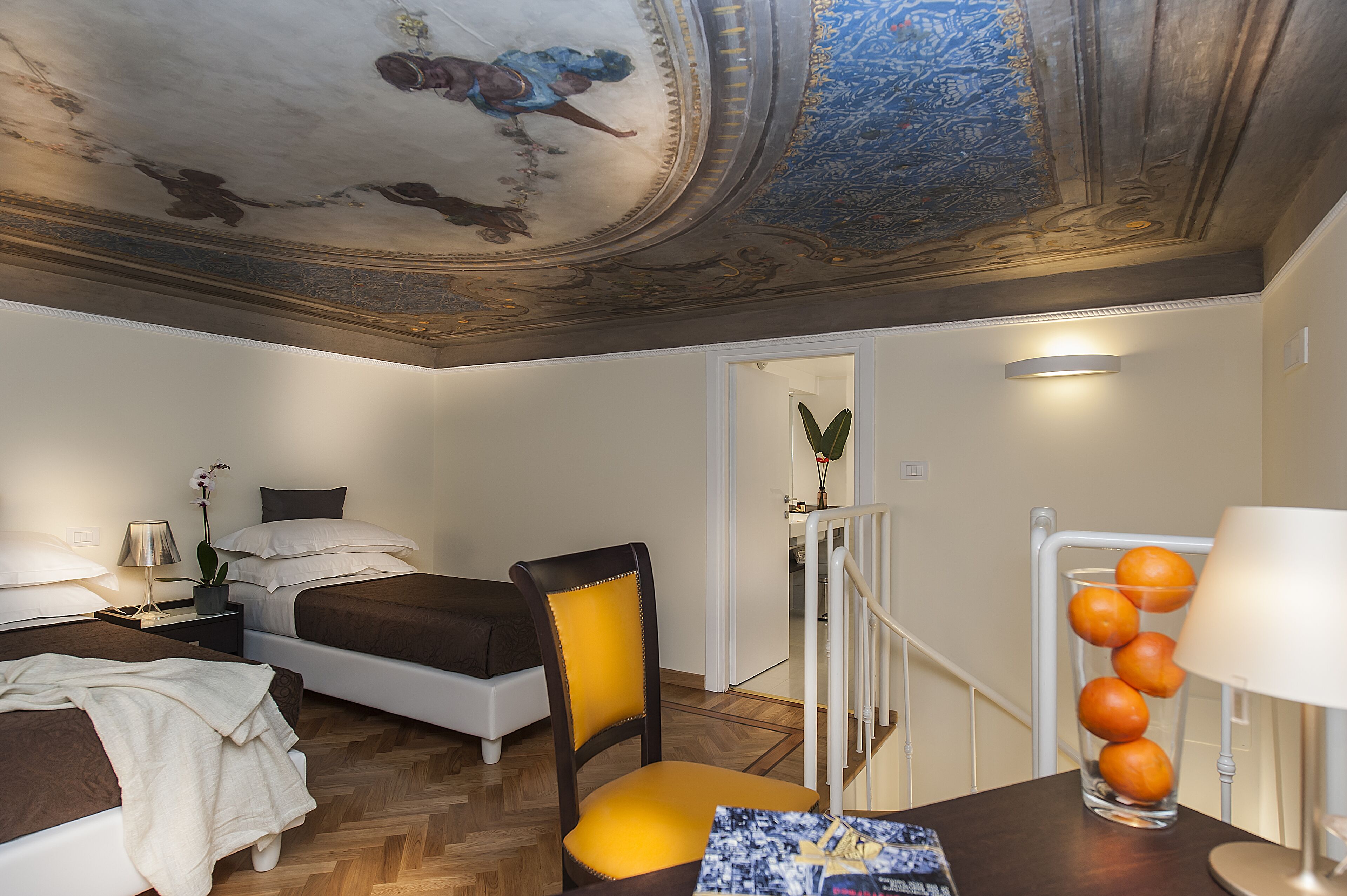Une chambre d’hôtel avec un lit, une chaise, un bureau avec une lampe, un vase rempli d’oranges et un plafond orné de motifs complexes.
