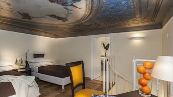 Une chambre d’hôtel avec un lit, une chaise, un bureau avec une lampe, un vase rempli d’oranges et un plafond orné de motifs complexes.