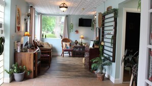 Ein gemütliches Wohnzimmer mit einem Holztisch, einer Couch und einem Sessel. Es befinden sich Blumentöpfe und ein Fernseher an der Wand.