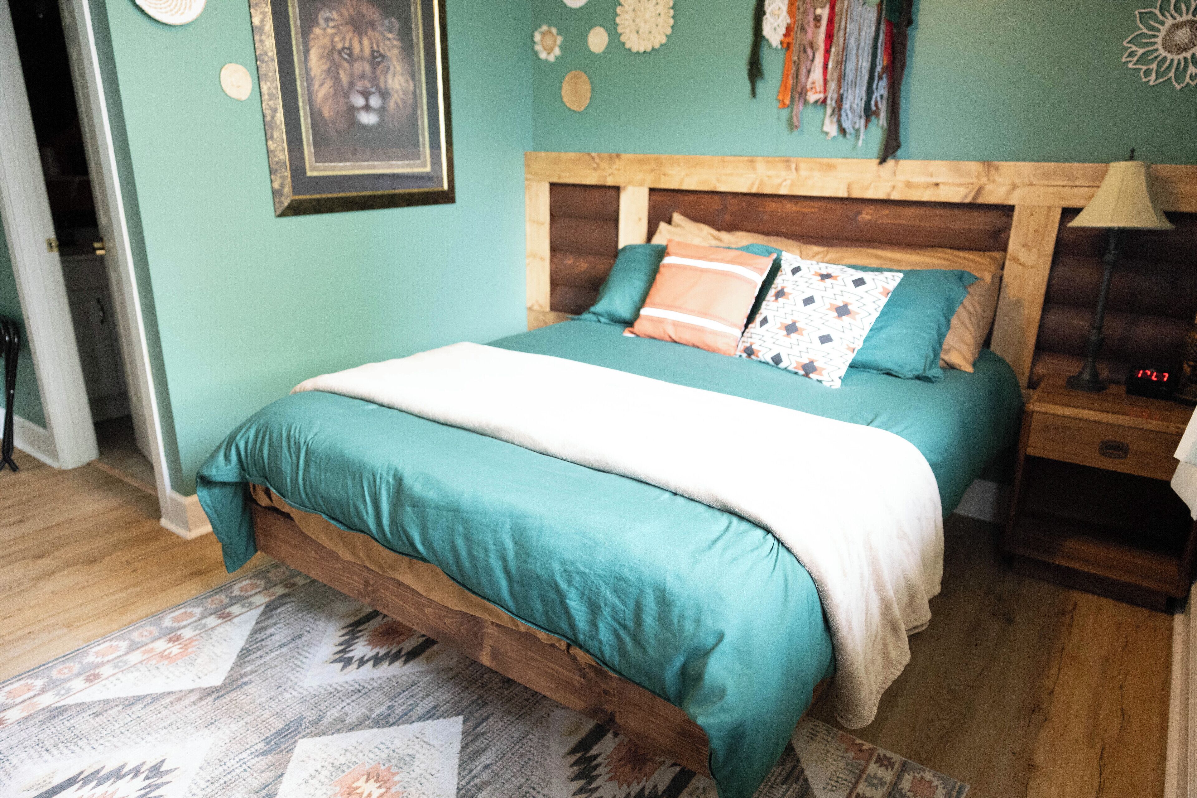 A bed with a wooden headboard, teal bedding, and patterned pillows. A patterned rug is on the floor, and a wall with decorative items is visible.