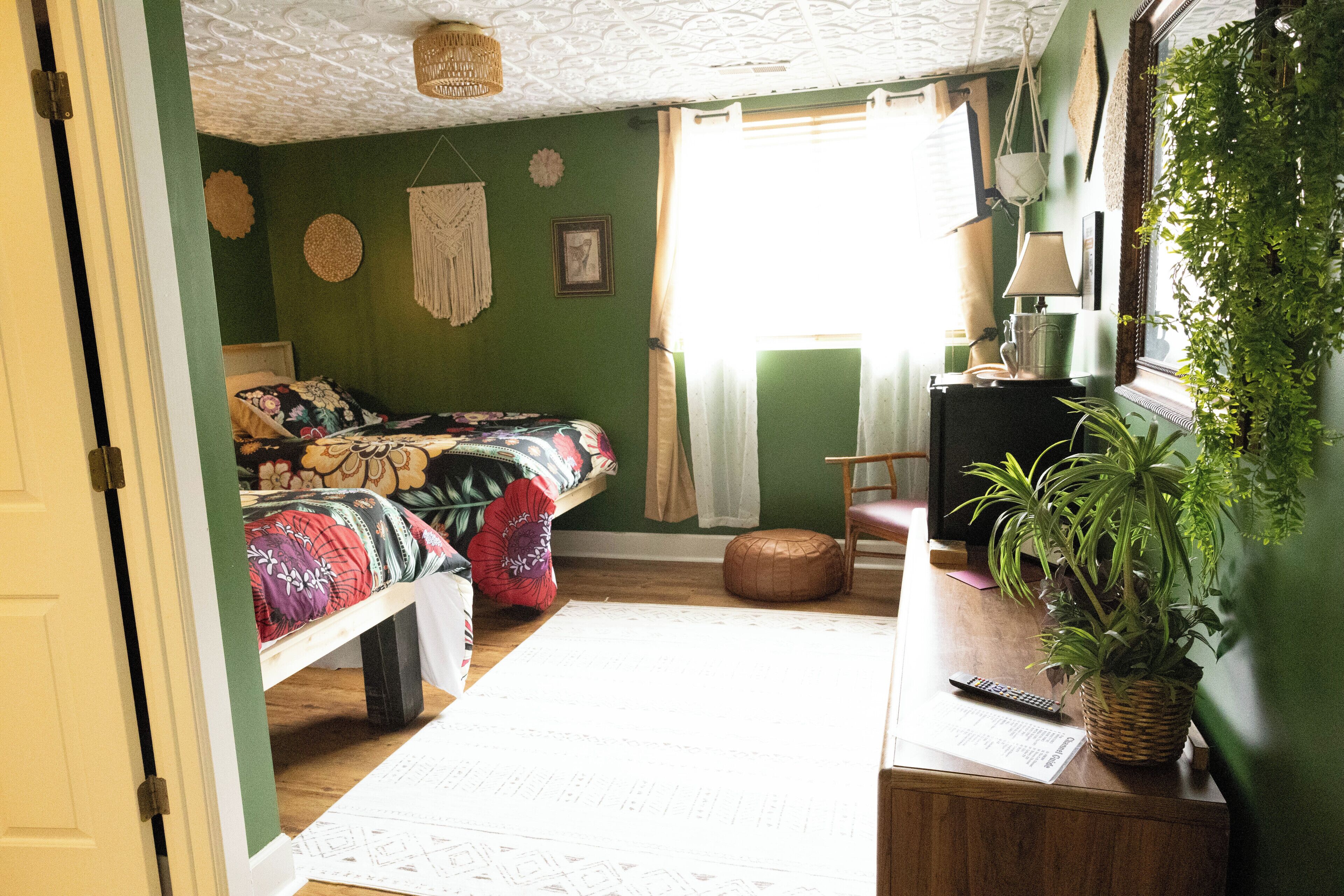 A room with a bed, a desk, a chair, a plant, and a window with curtains.