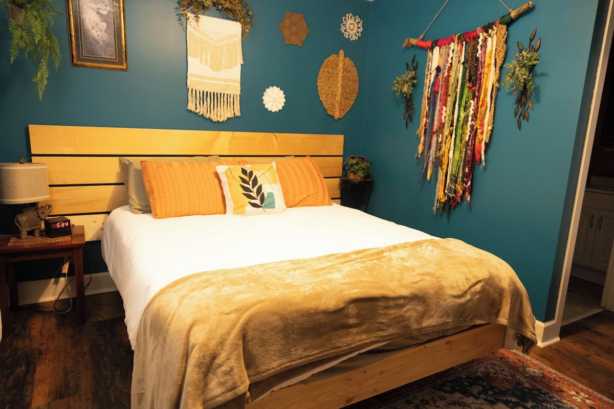 A bedroom with a wooden bed, a nightstand with a lamp, and a wall decorated with textiles and wall hangings.