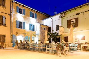 Un café en plein air, avec des tables et des chaises, entouré d’immeubles aux volets bleus, et un vélo garé à proximité.